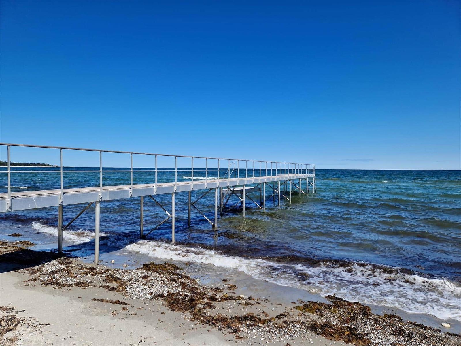 Ein langer Steg führt ins Meer. Strand und tiefblaues Wasser sind sichtbar.