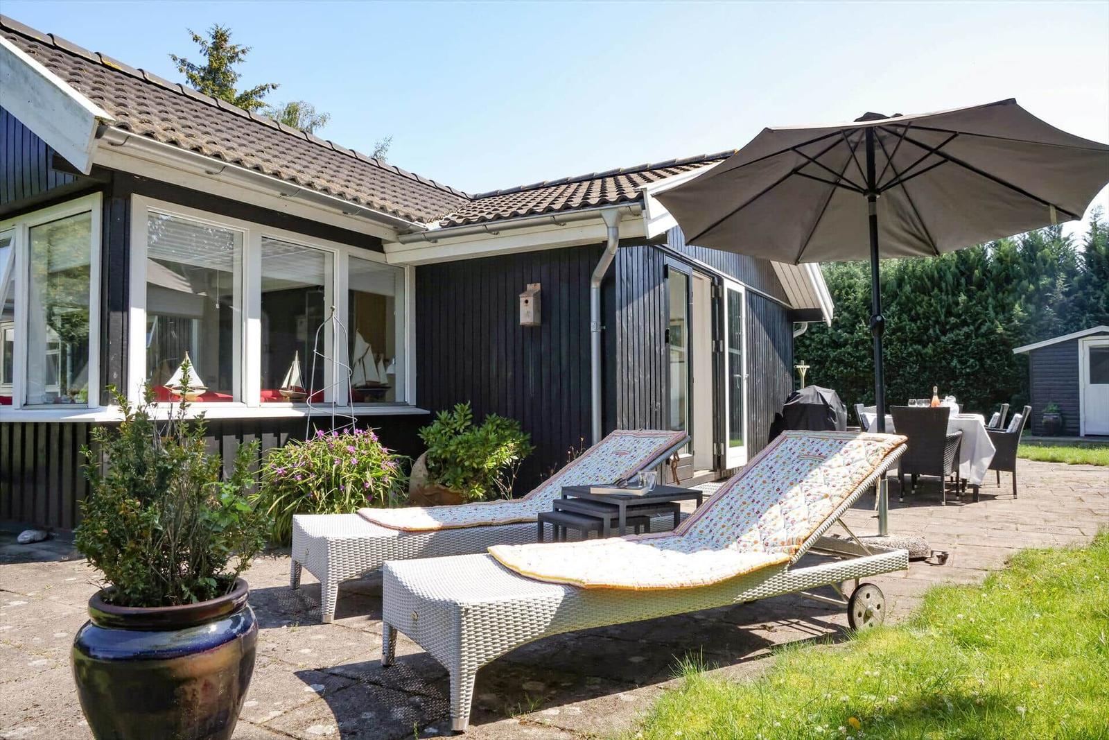 Terrasse mit Liegestühlen, Sonnenschirm und Gartenmöbeln vor einem Haus mit dunkler Fassade.