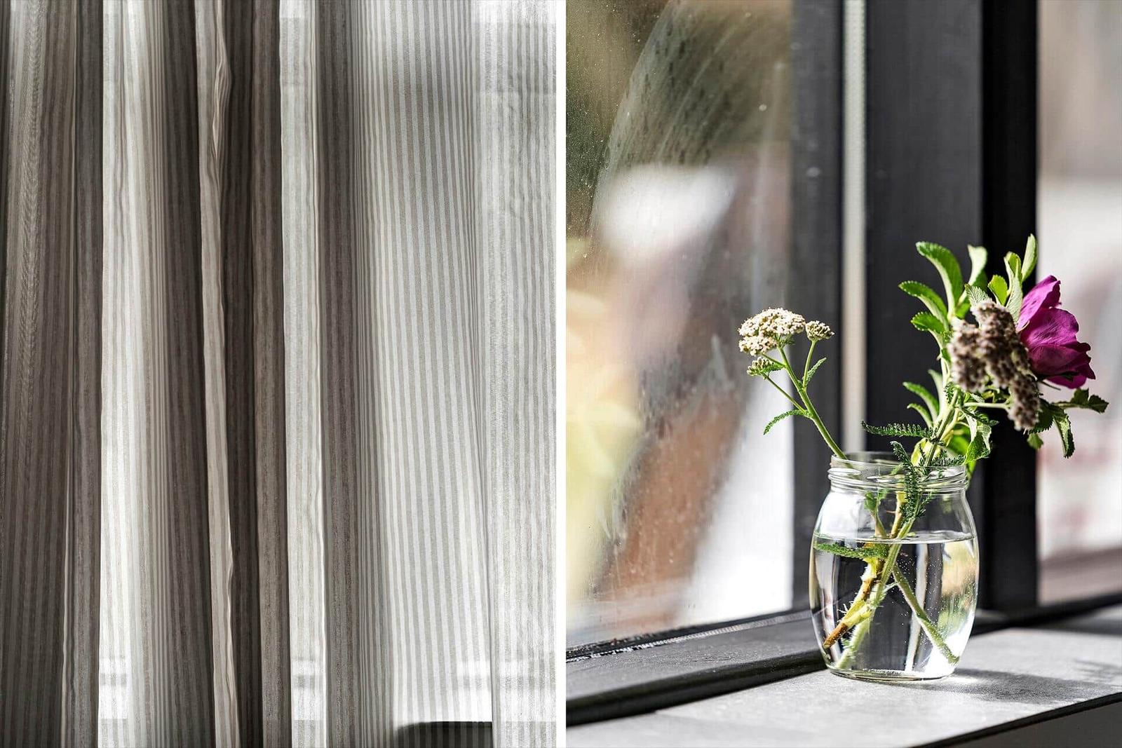 Glasvase mit Blumen auf Fensterbank neben markanten Vorhängen.