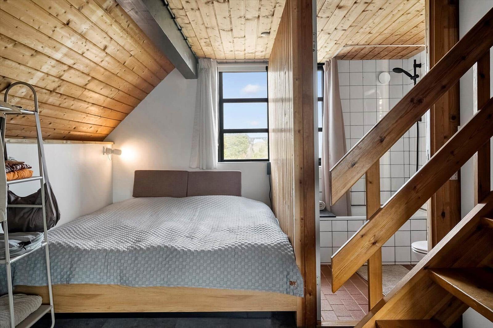 Schlafzimmer mit Dachschrägen, Fenster und Holztreppe zur Toilette.