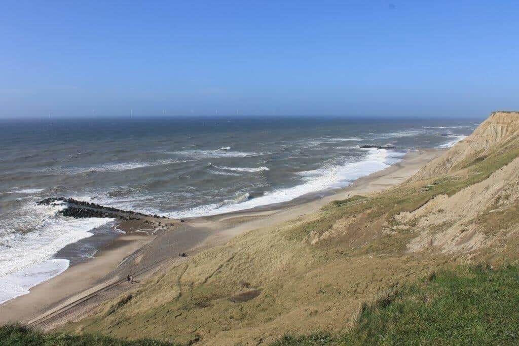Strand mit Dünen und Wellen, Blick auf das Meer und Windkraftanlagen im Hintergrund.