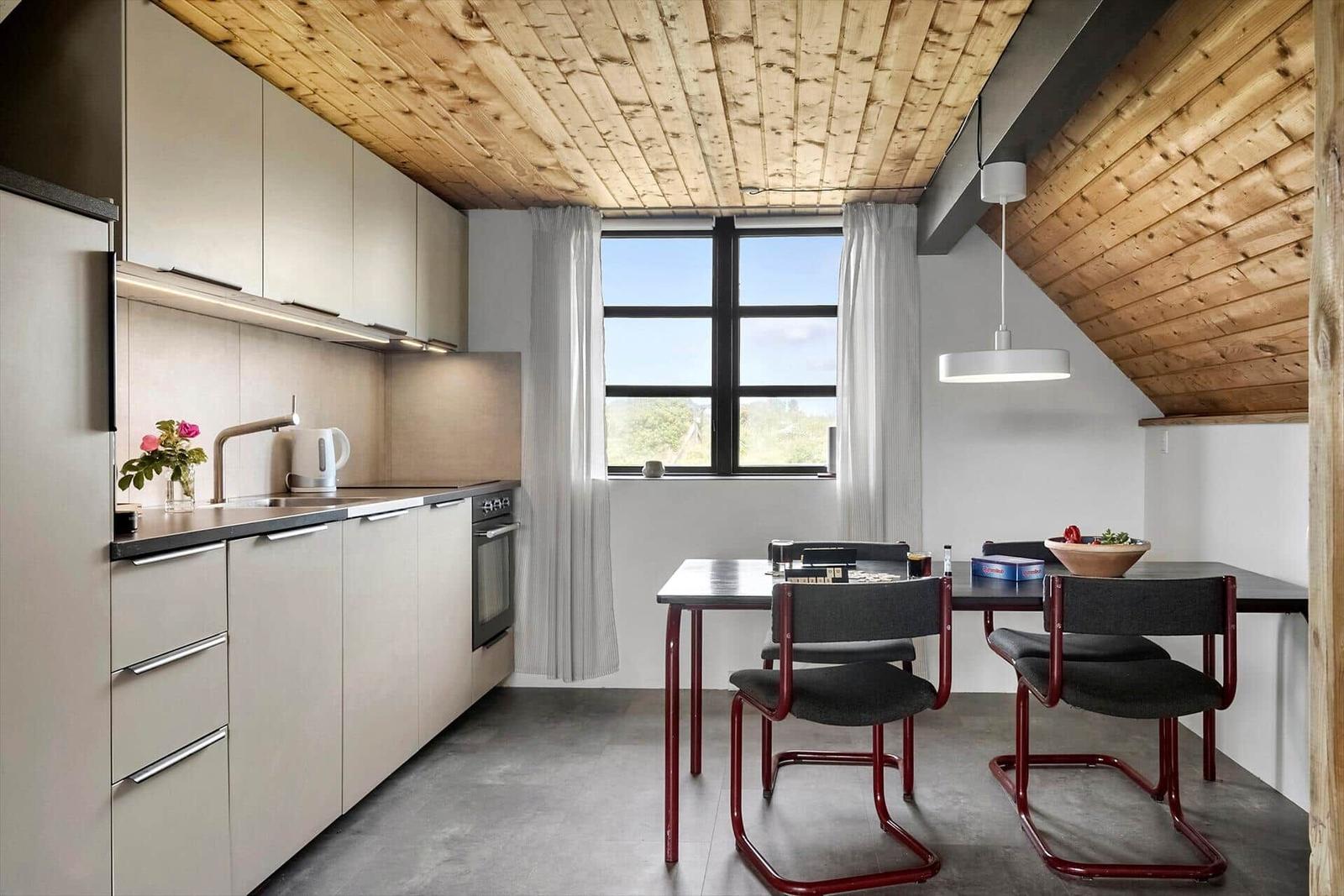 Küche mit Essbereich unter schrägem Holzdach. Fenster, Tisch, Stühle und Küchenschränke.