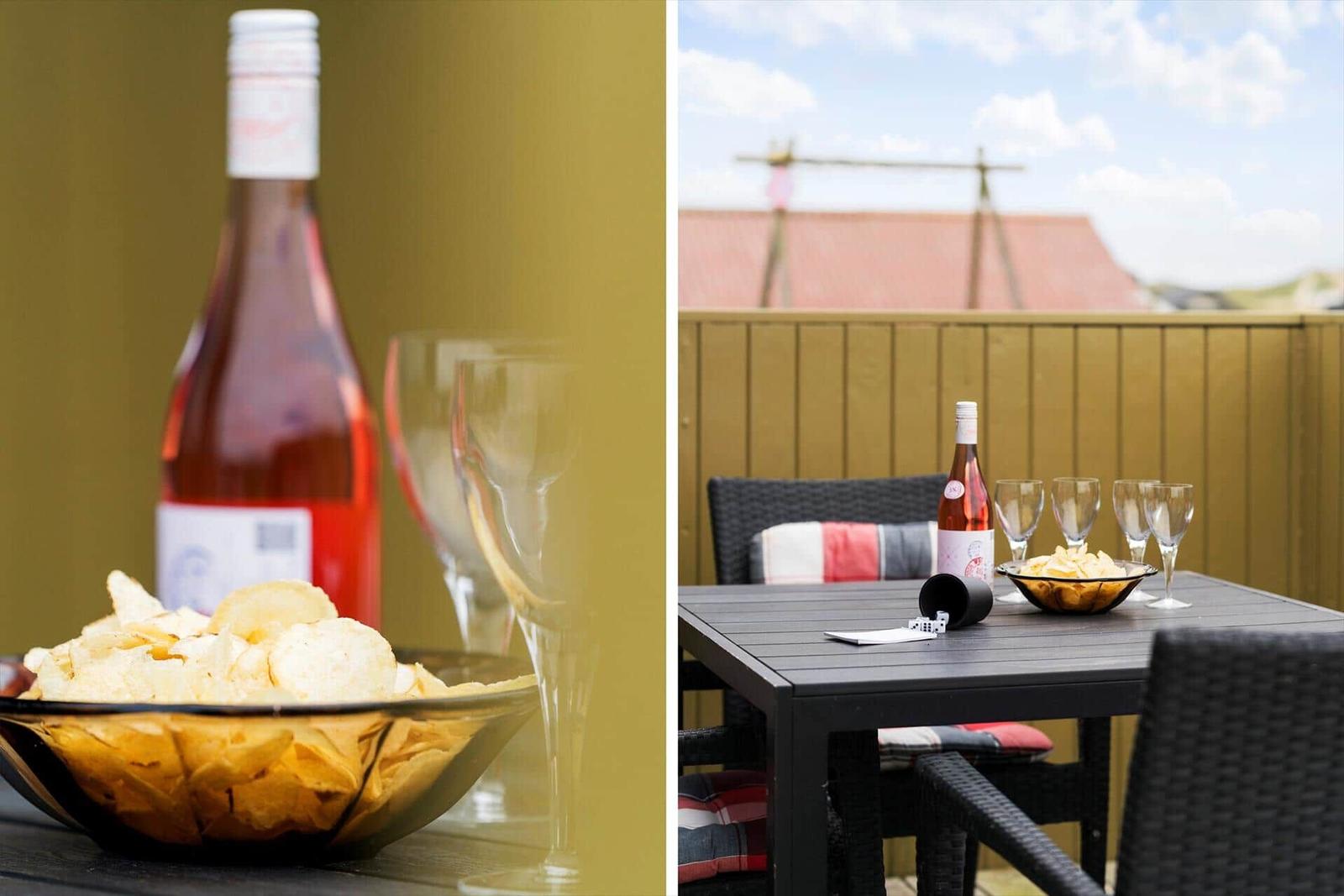 Terrasse mit Tisch, Stühlen, Wein und Chips. Hintergrund: Holzzaun und Dach.