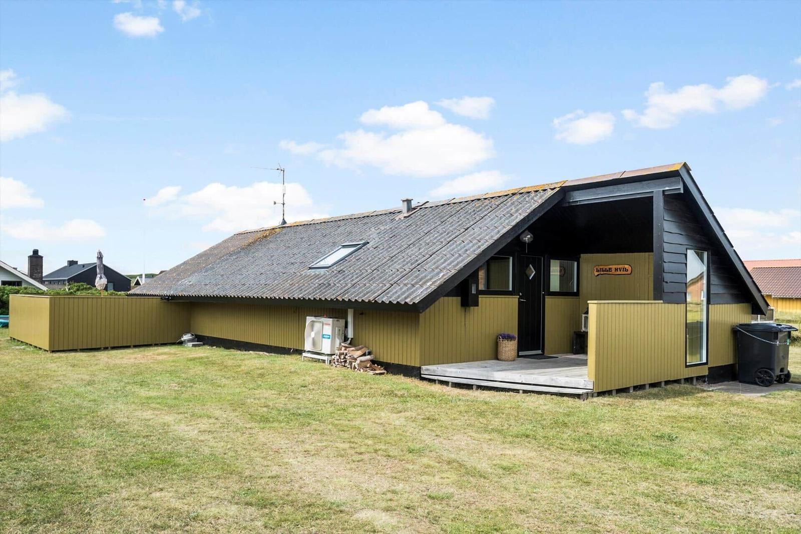 Holzhaus mit grünem Zaun und Holzstapel im Garten unter blauem Himmel.
