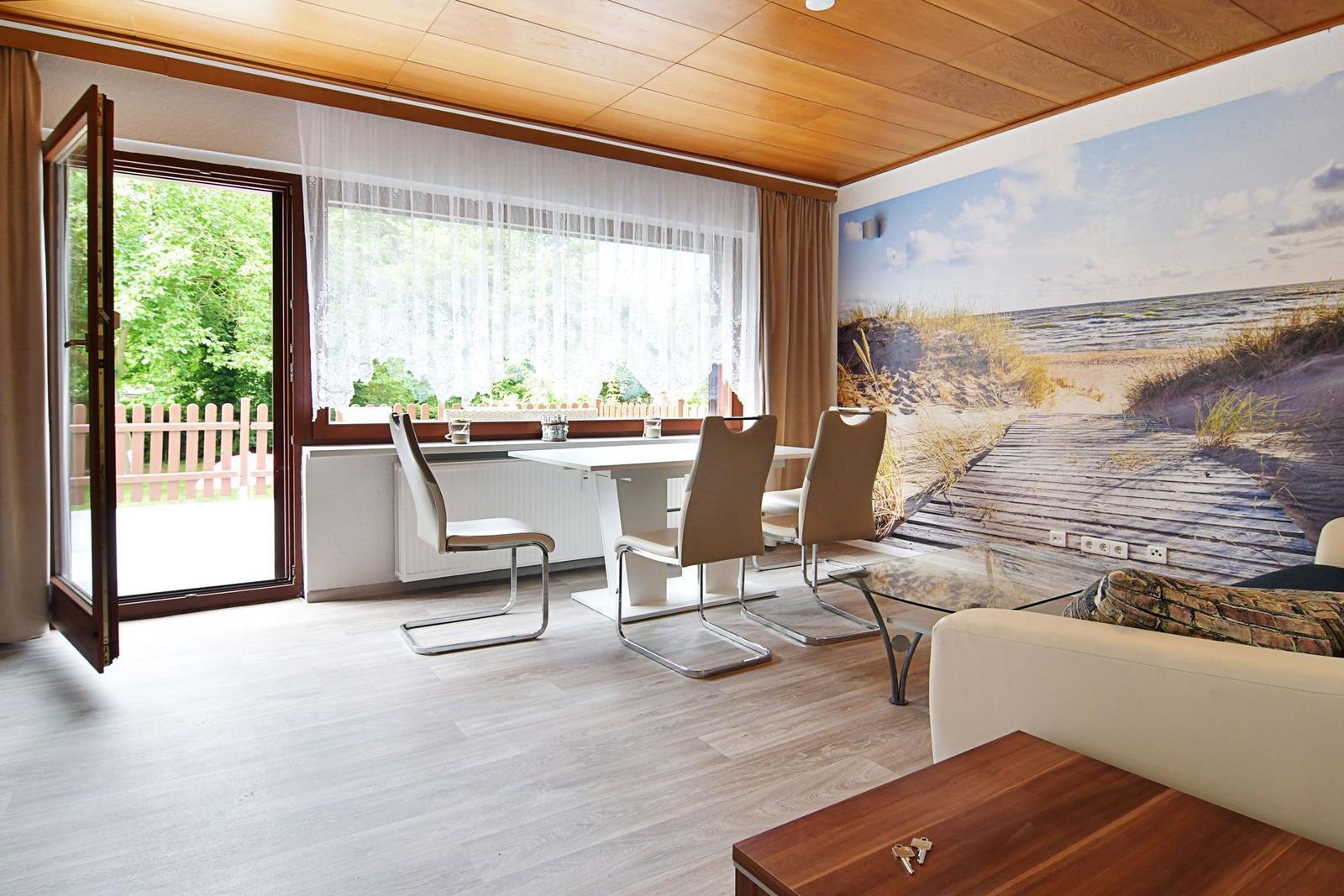 Esszimmer mit Tisch, Stühlen und Blick auf Garten. Wandbild zeigt Strand.