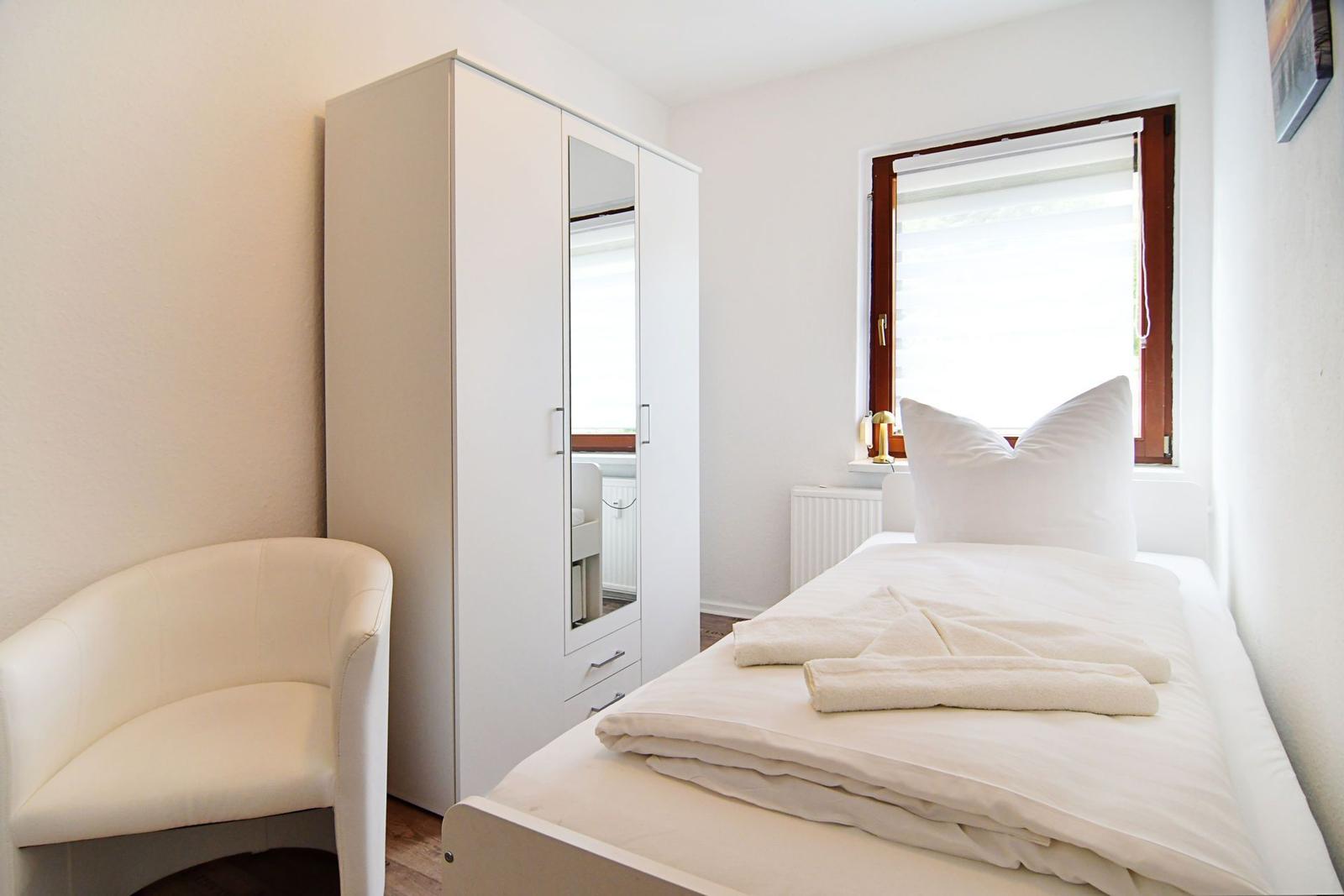 Ein Schlafzimmer mit weißem Bett, Kleiderschrank und Sessel. Fenster mit Jalousie.