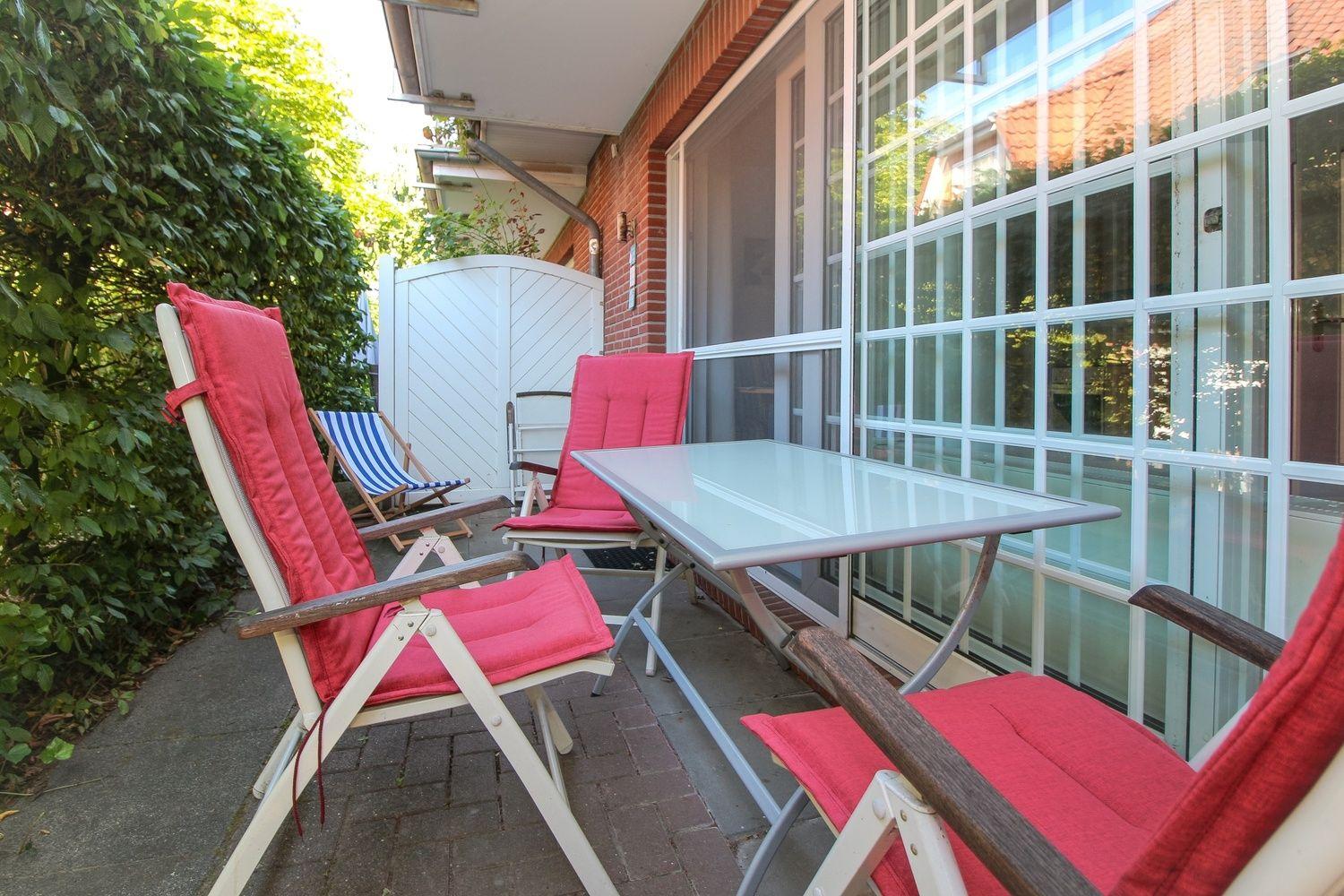 Terrasse mit Tisch und drei Stühlen mit roten Kissen. Hintergrund: Glasfront und grüne Hecke.
