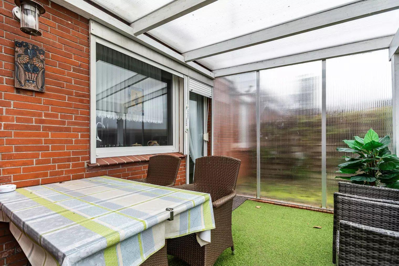 Terrasse mit Tisch, Stühlen und künstlichem Gras unter transparentem Dach.