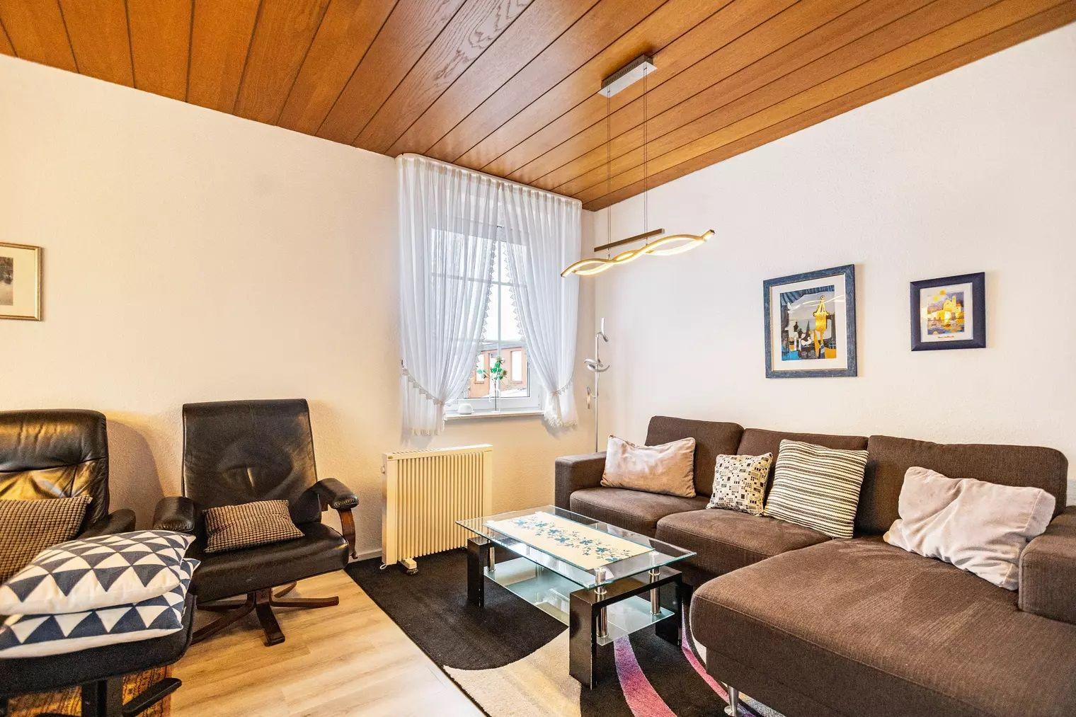 Wohnzimmer mit Sofa, Sessel, Glas-Tisch und Holzdecke.