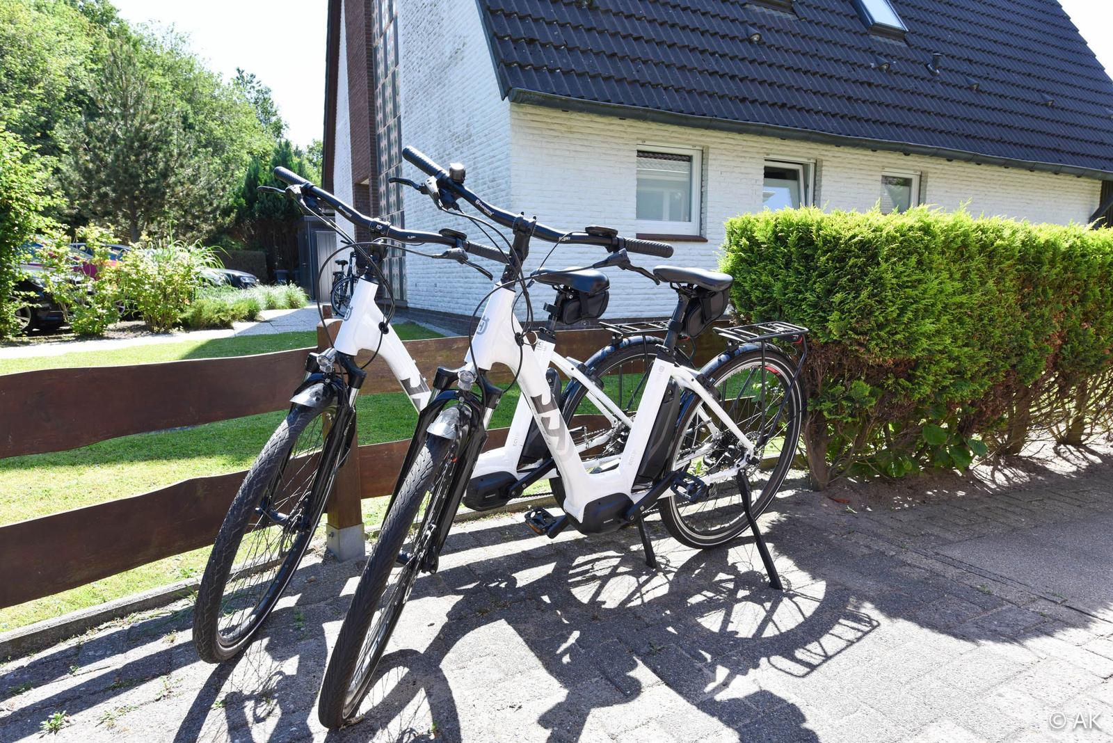 Zwei weiße E-Bikes stehen vor einem Haus mit grünem Heck.