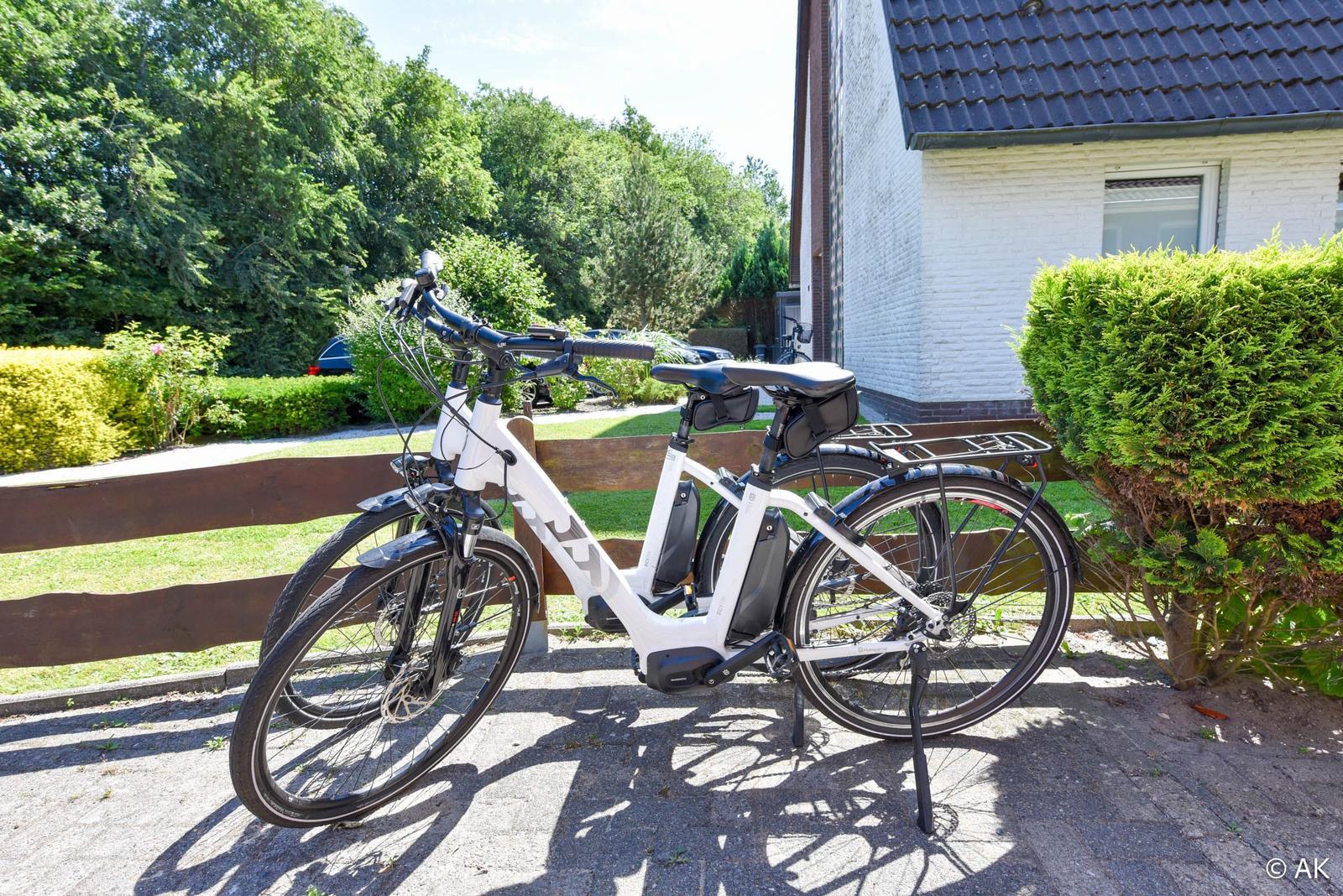 Ein weißes E-Bike steht vor einem Haus mit Garten und Hecke.