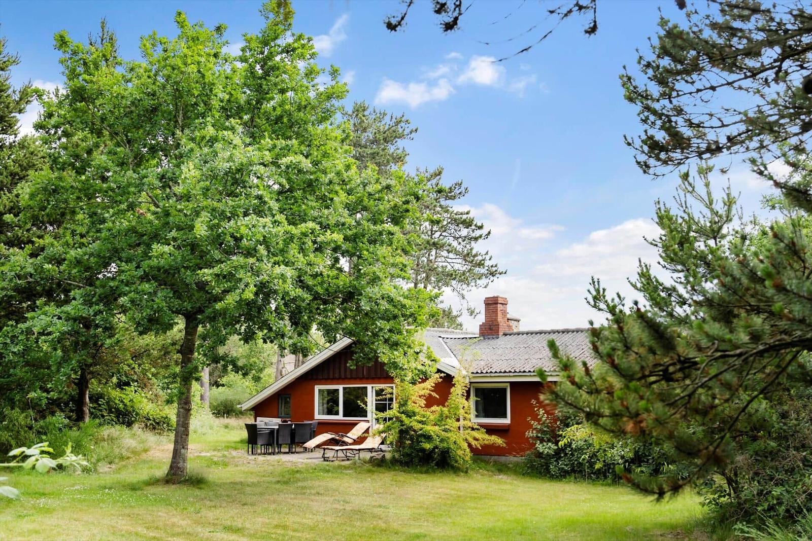 Rotes Haus mit Garten, Terrasse und Gartenmöbeln unter Bäumen.