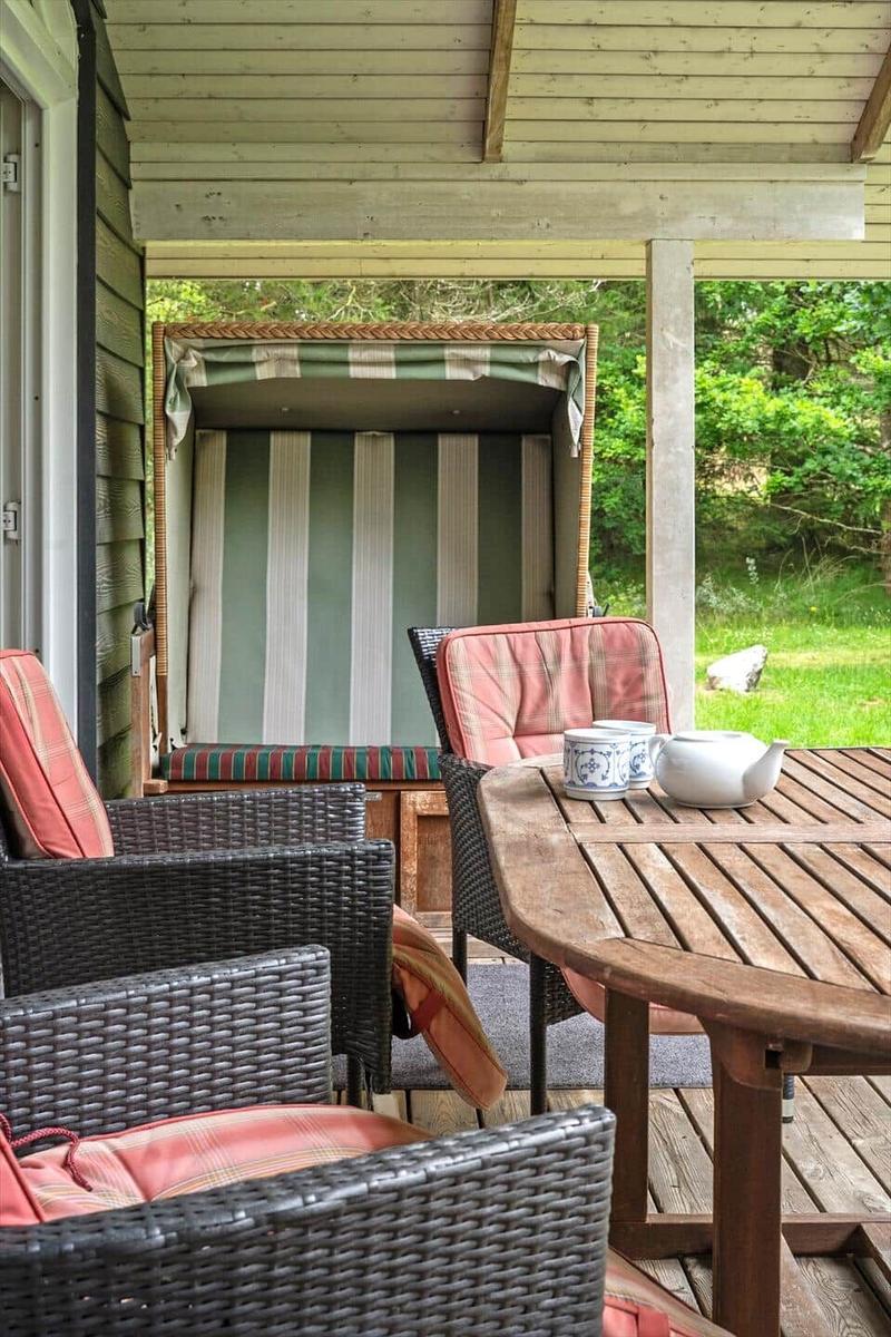 Terrasse mit Holztisch, Wickerstühlen und Strandkorb. Grün hinter dem Haus.