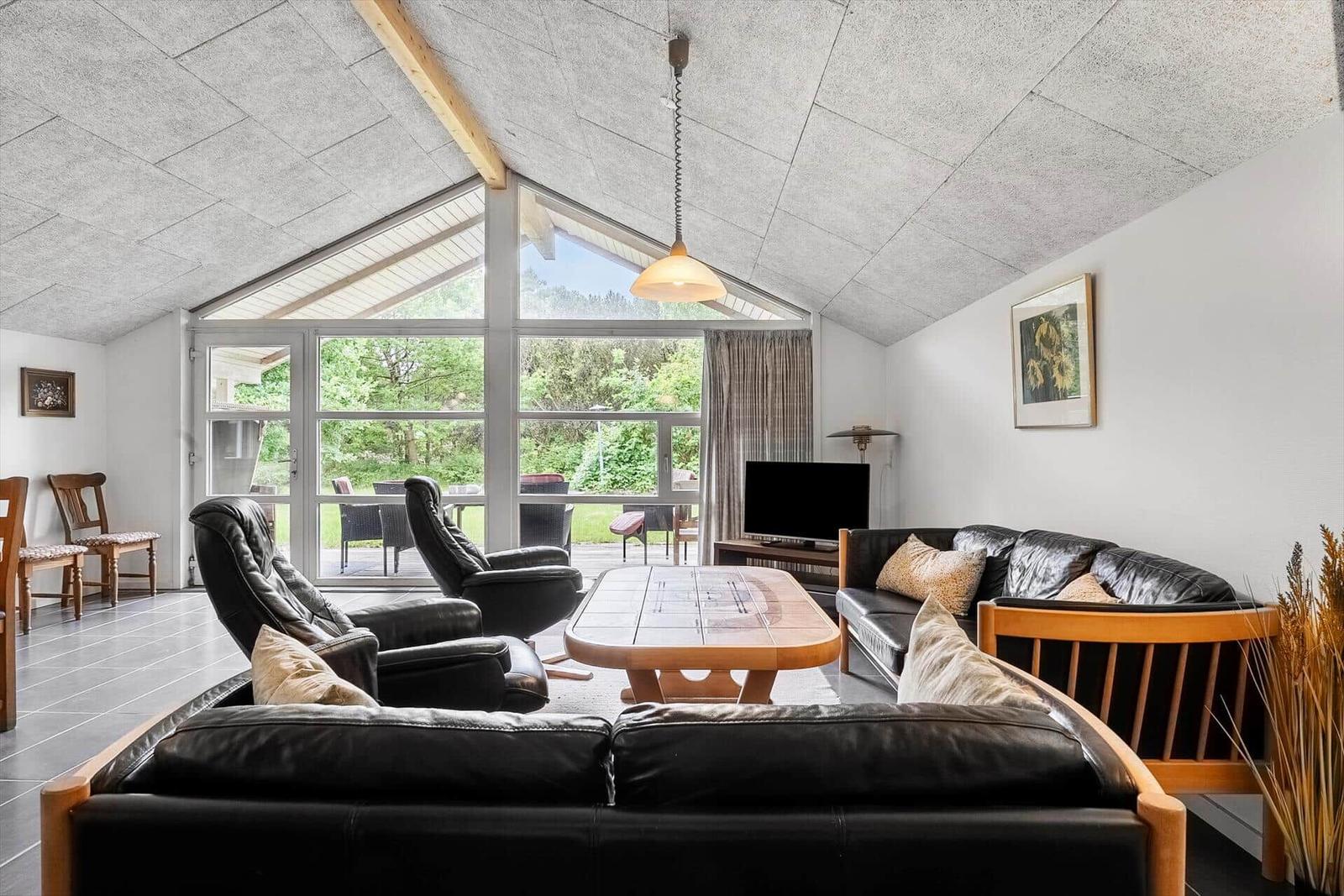 Wohnzimmer mit Sofa, Sesseln und TV. Große Fensterfront mit Blick auf Garten.