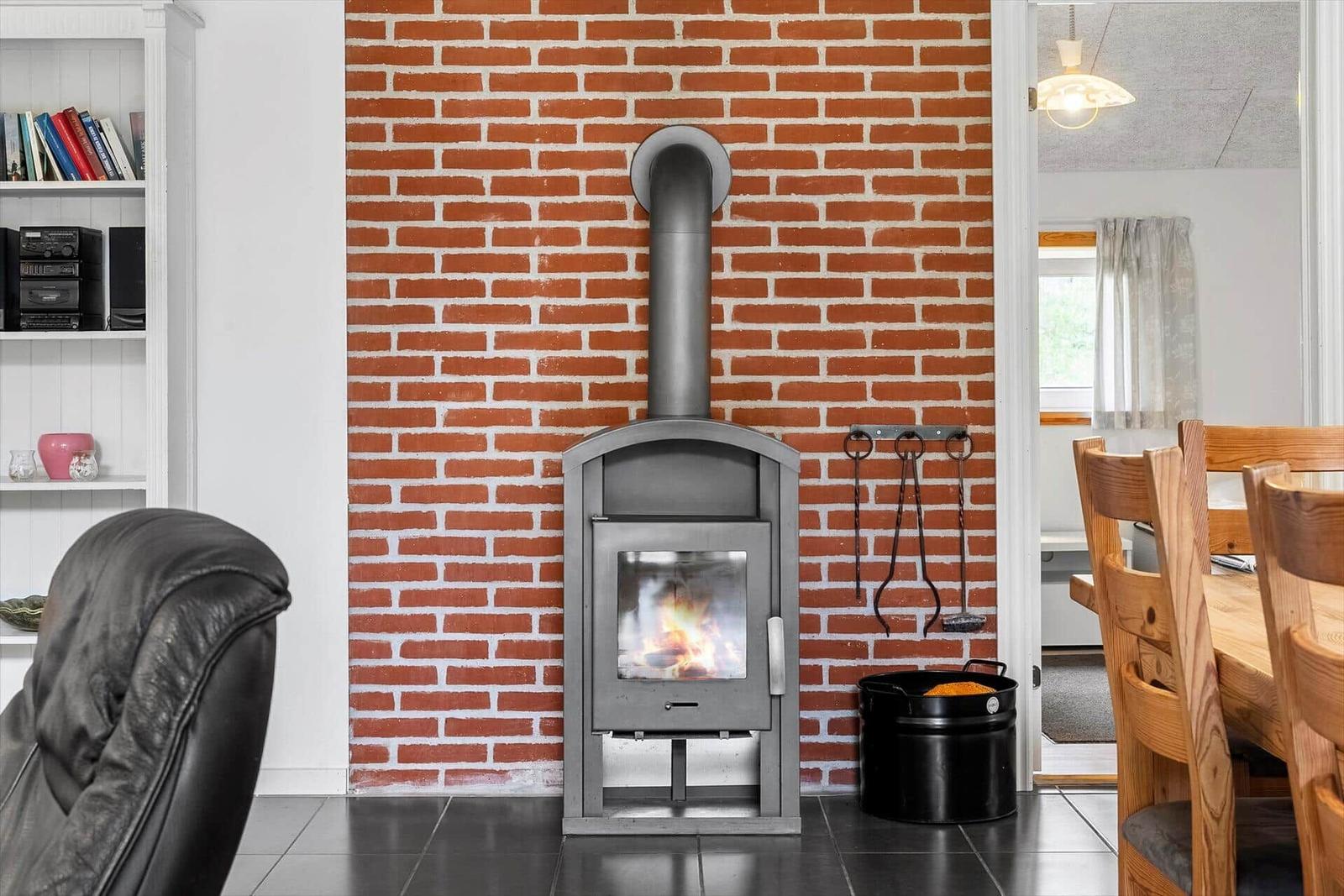 Ein Kaminofen steht vor einer Backsteinwand. Links ein Bücherregal, rechts ein Holztisch mit Stühlen.