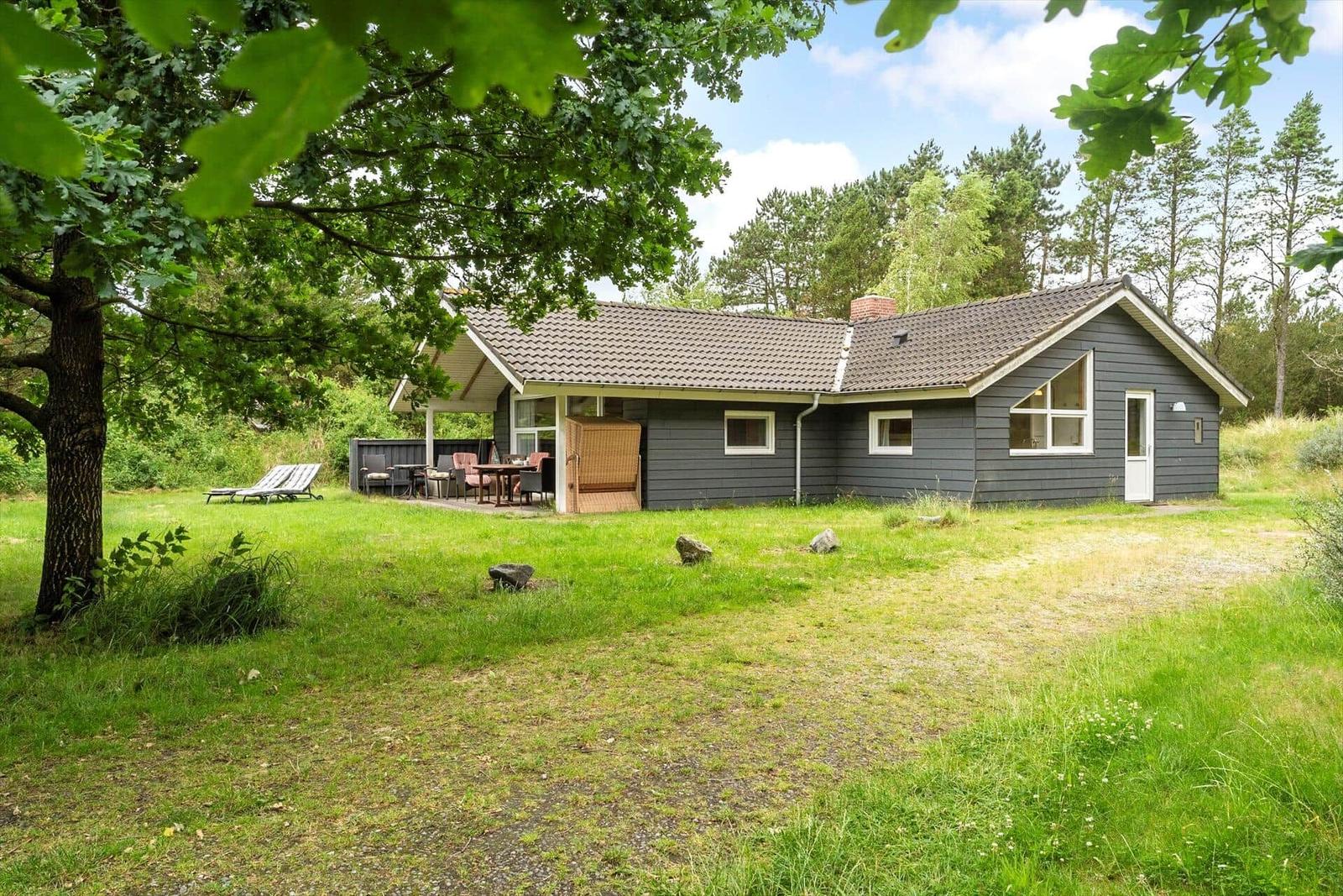 Ein graues Ferienhaus mit Terrasse und Garten unter Bäumen.