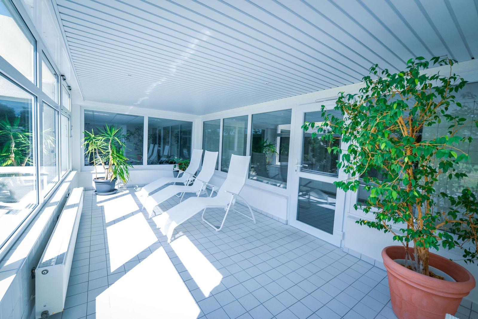 Terrasse mit Liegestühlen, Pflanzen und Glaswänden. Fliesenboden und weißer Dachbalken.