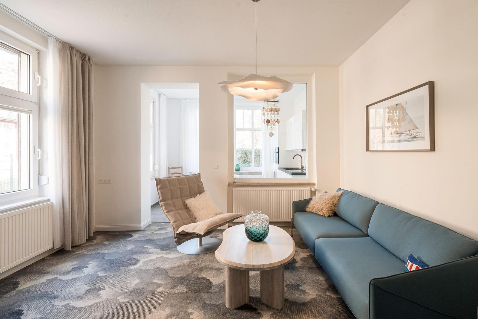 Wohnzimmer mit Sofa, Sessel und Tisch. Küche im Hintergrund sichtbar.