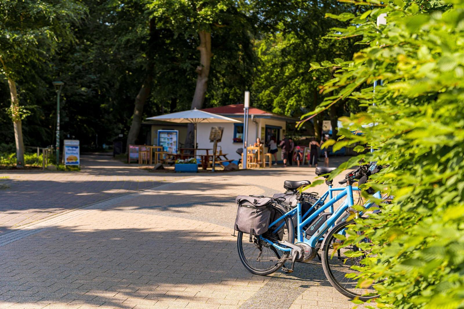 Ein blauer E-Bike steht vor einem Restaurant im Park. Menschen sitzen draußen.