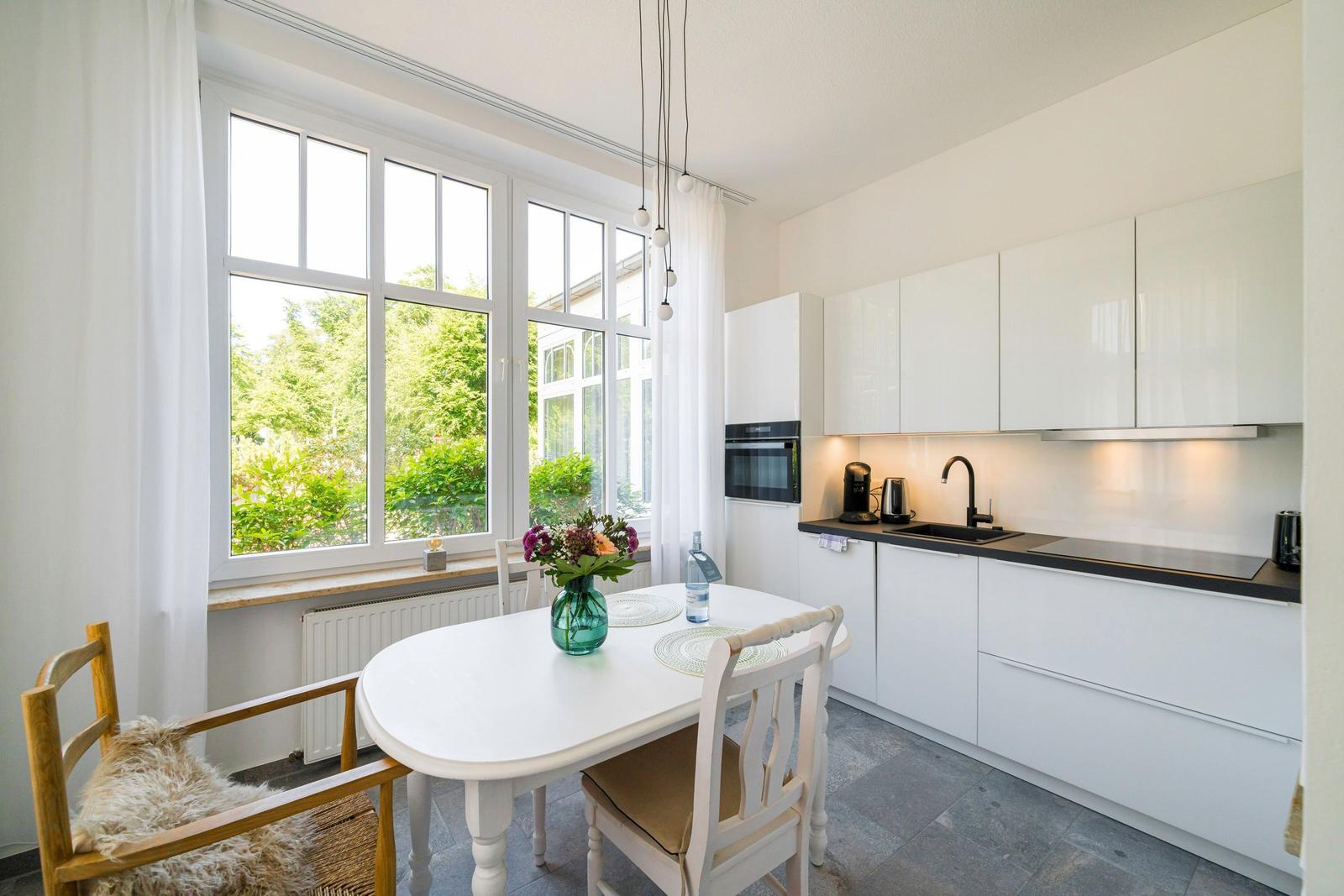 Küche mit Esstisch, großen Fenstern und weißen Schränken.