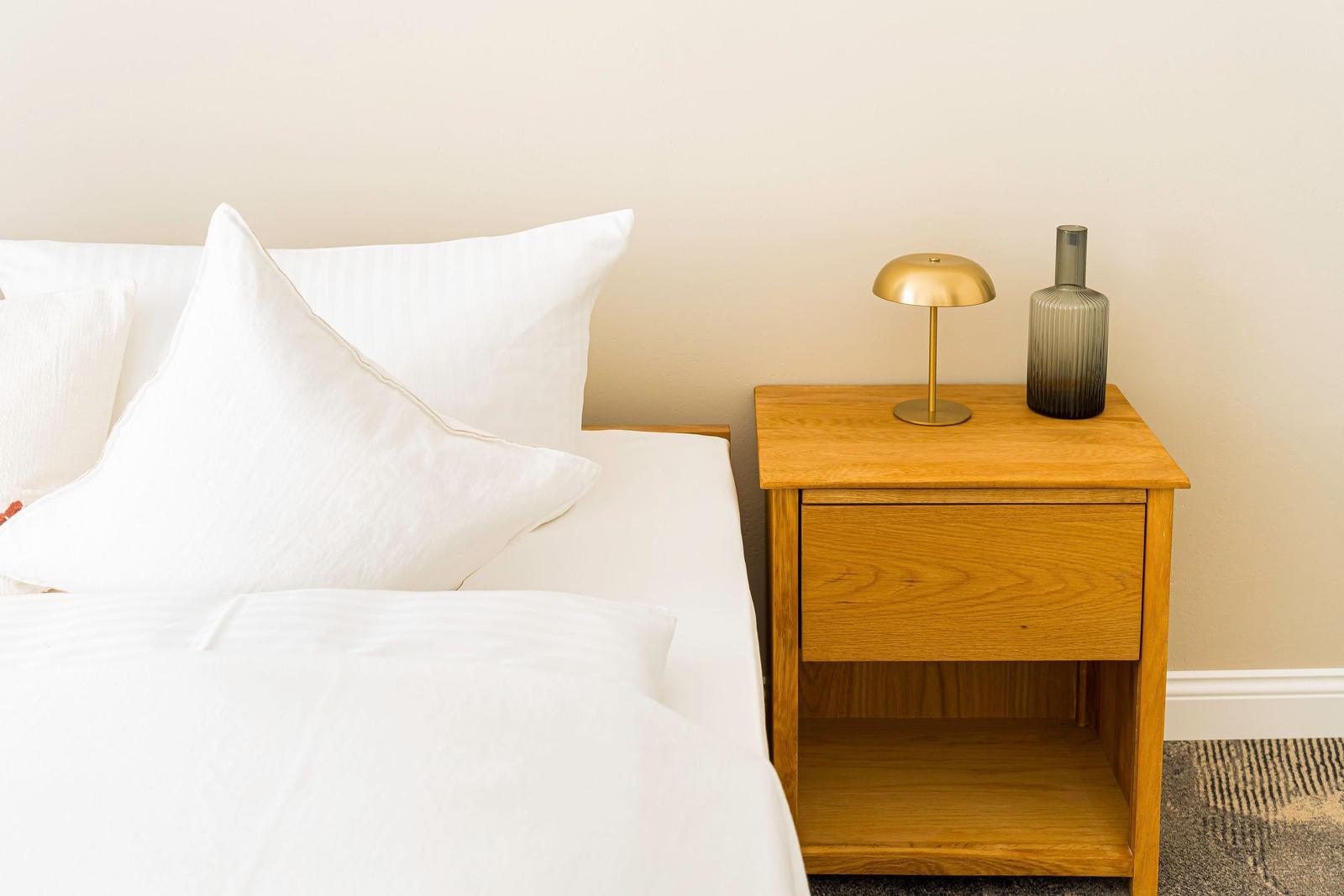 Ein Bett mit weißen Kissen und einer Nachttischlampe neben einem Holznachttisch.