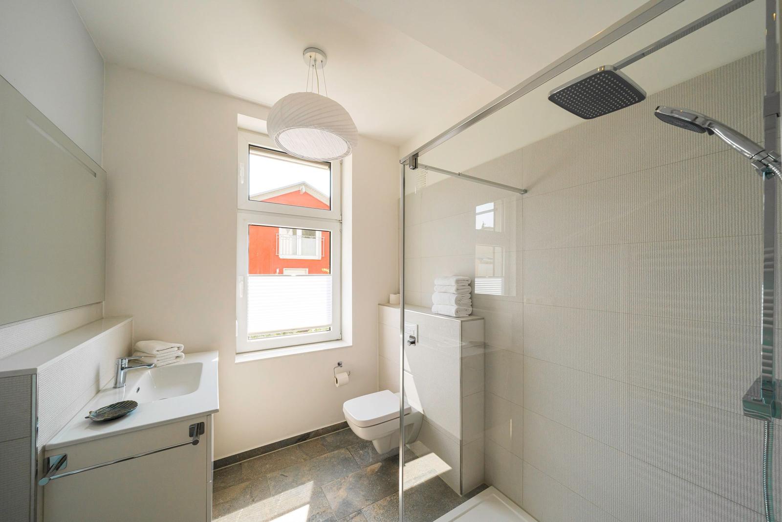 Bad mit Dusche, Waschbecken und Toilette. Fenster mit Blick auf rotes Gebäude.