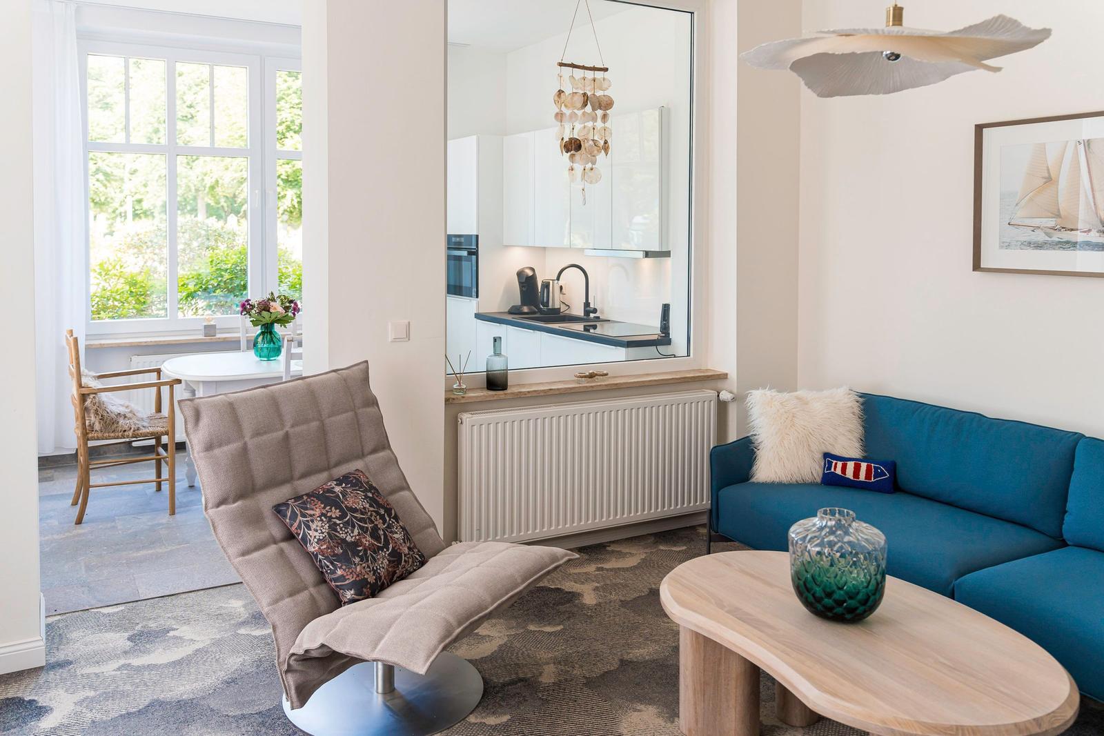 Wohnzimmer mit blauem Sofa, Holztisch und Sessel. Küche im Hintergrund über Glaswand.