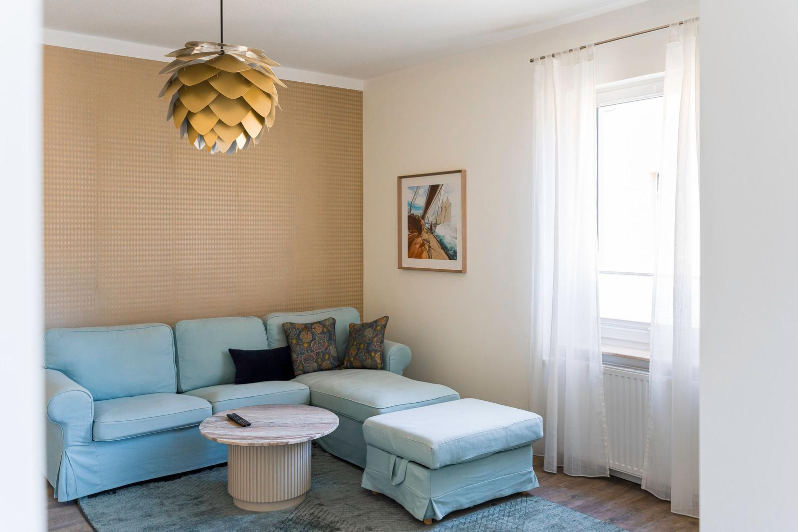 Ein Wohnzimmer mit Sofa, Couchtisch und Fenster. Deckenleuchte und Wandbild sind sichtbar.