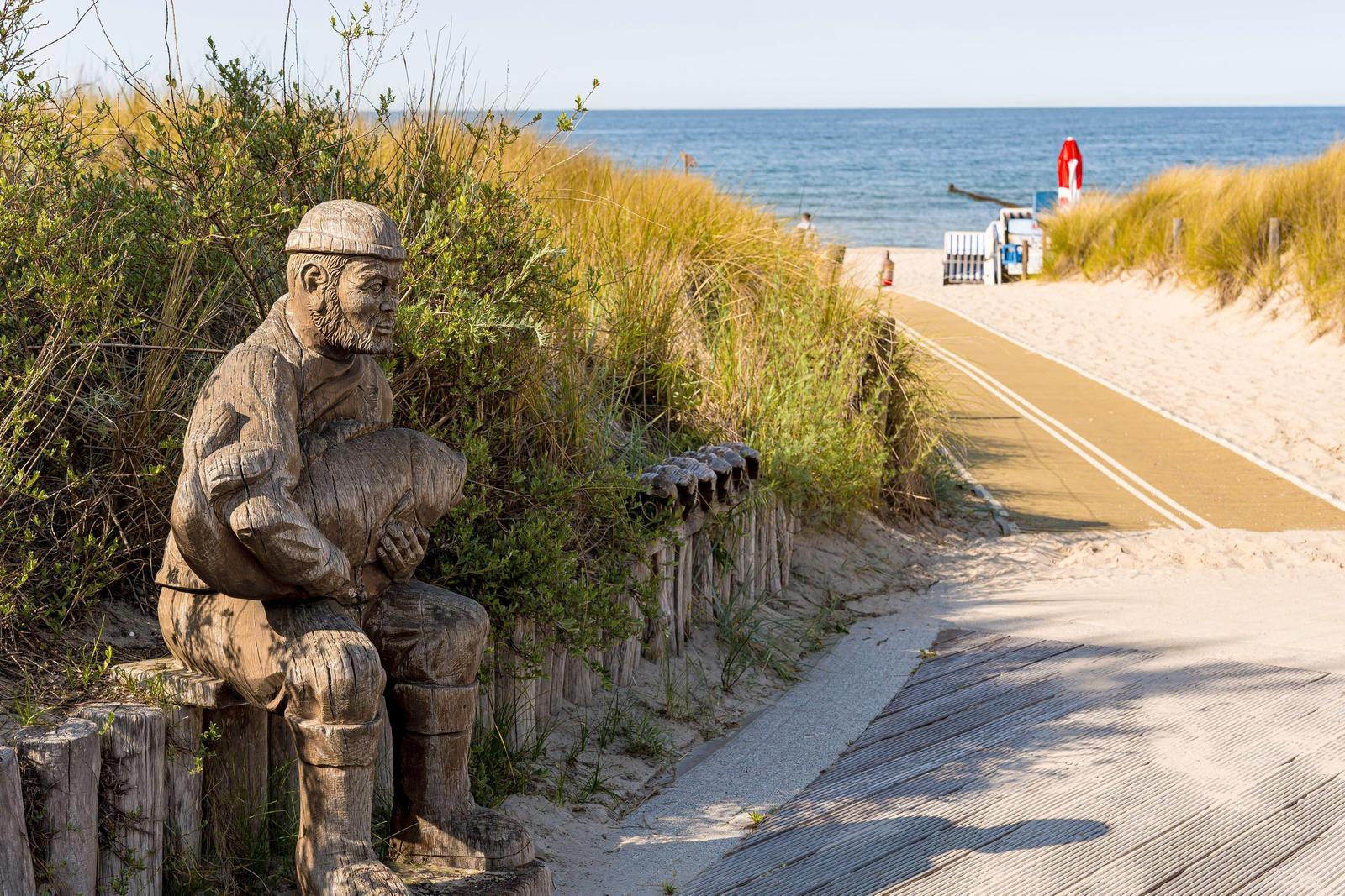 Holzskulptur eines Fischer neben Strandweg mit Blick auf Meer.