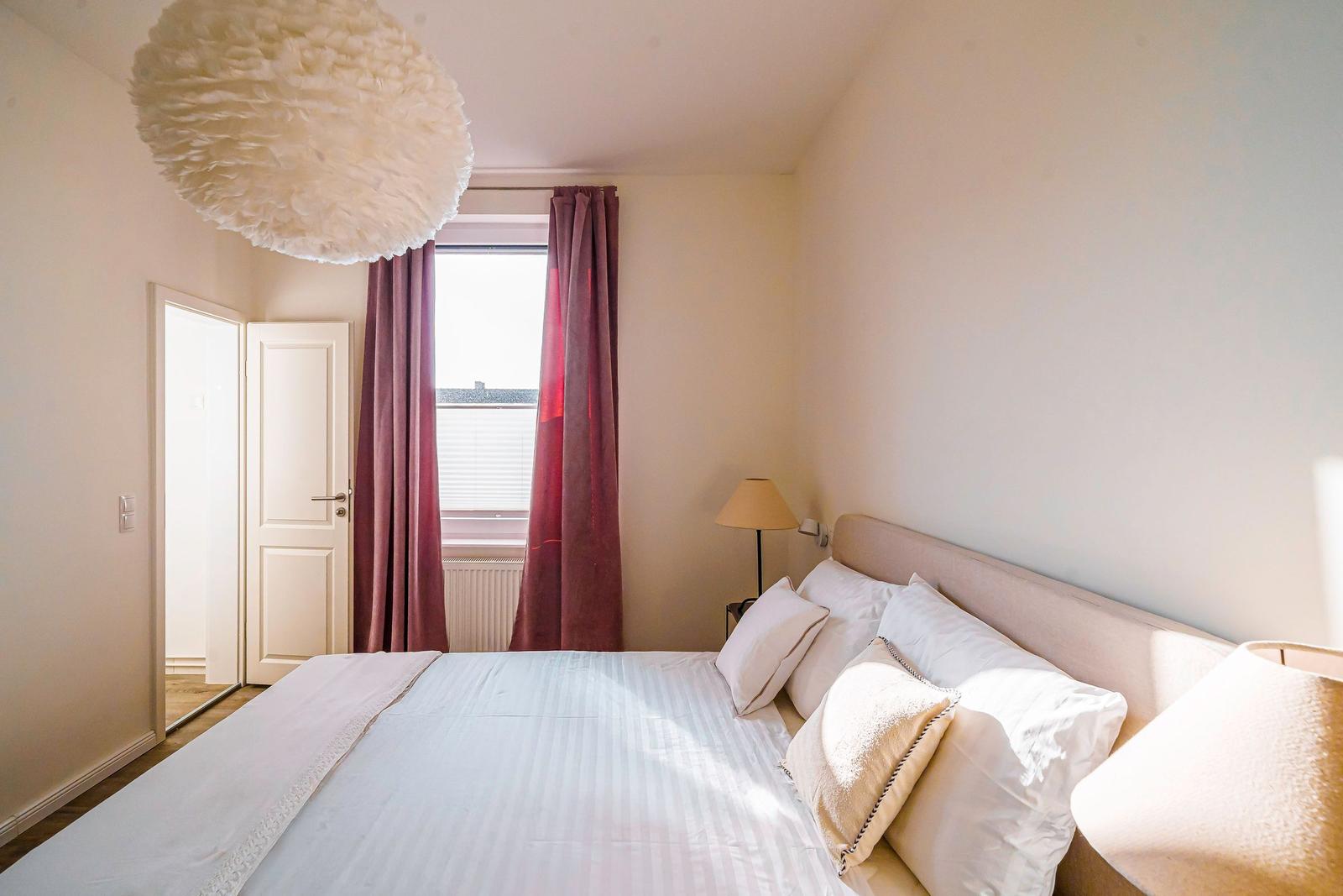 Schlafzimmer mit Bett, Fenster, Vorhängen und Deckenleuchte.