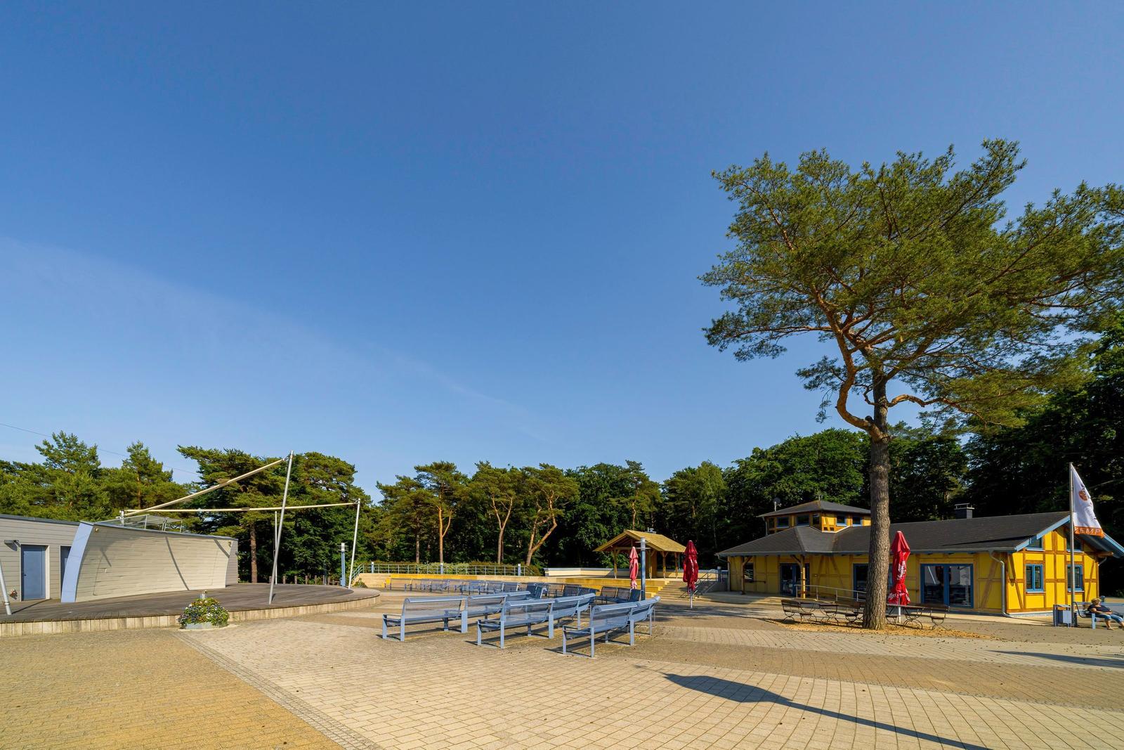 Freiluftbühne mit Bänken, gelbes Gebäude und großer Baum unter blauem Himmel.