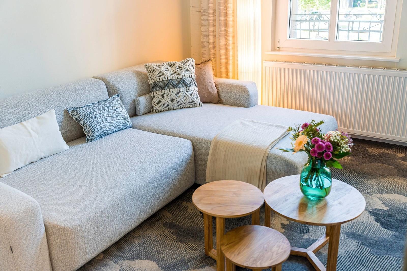 Ein Sofa mit Kissen und zwei runden Tischen. Ein Fenster und ein Heizkörper sind sichtbar.