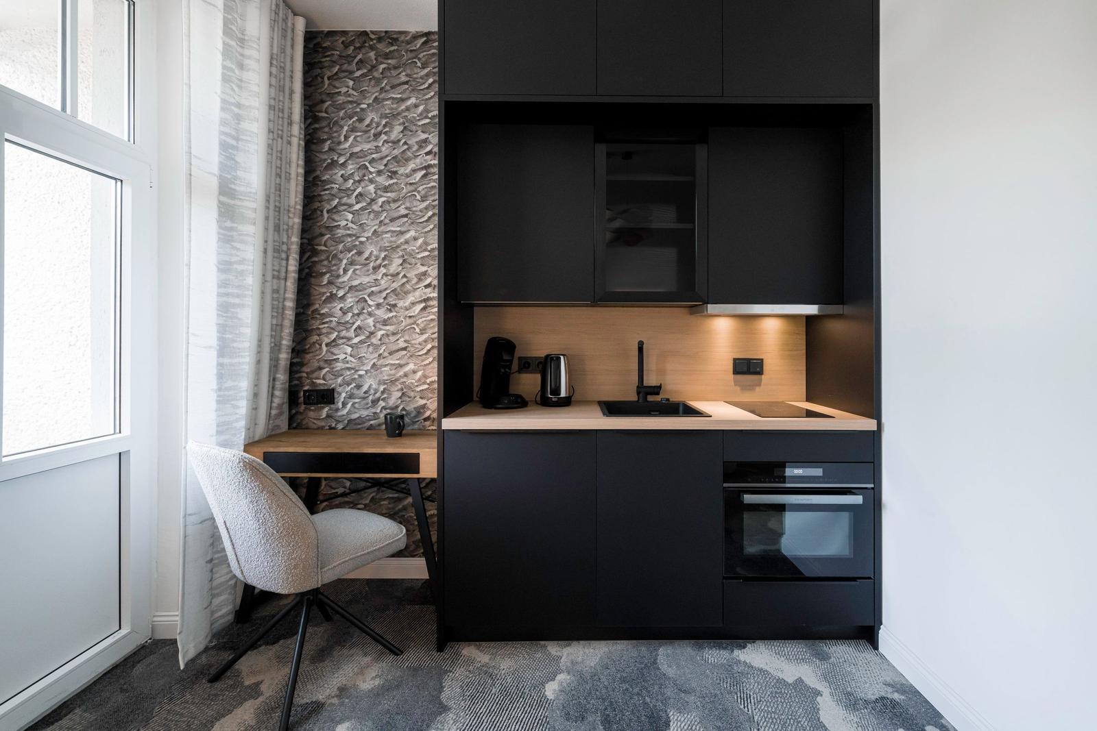 Küche mit Arbeitsfläche, Spüle und Ofen. Nebenher Schreibtisch mit Stuhl und Fenster.