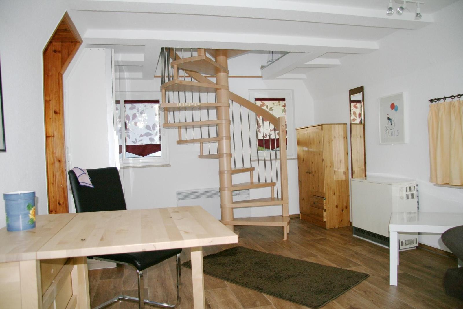 Raum mit Holztreppe, Tisch, Stuhl und Kleiderschrank. Fenster mit Vorhängen.