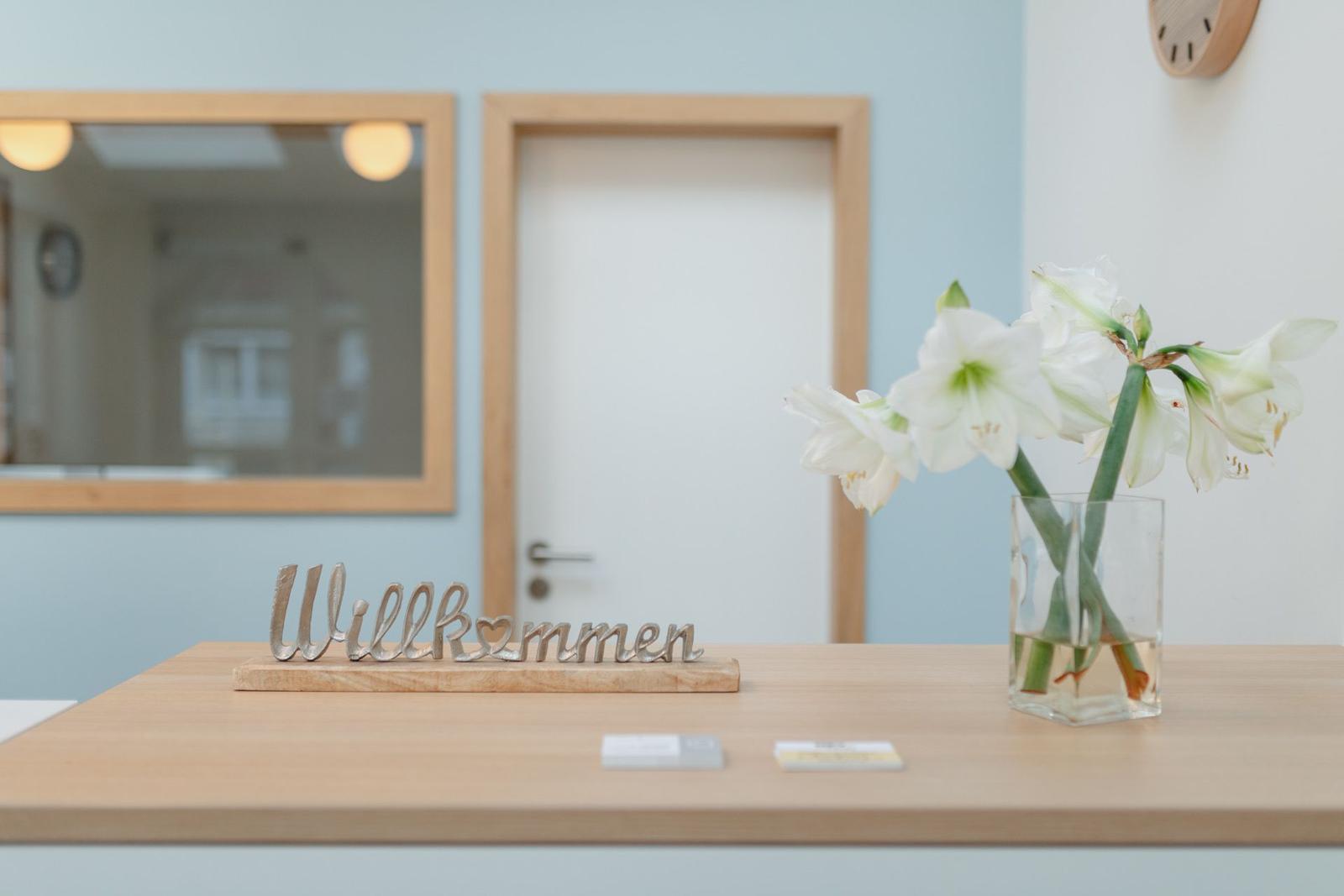 Willkommen-Schild und Blumen auf Holztisch vor Tür und Spiegel.