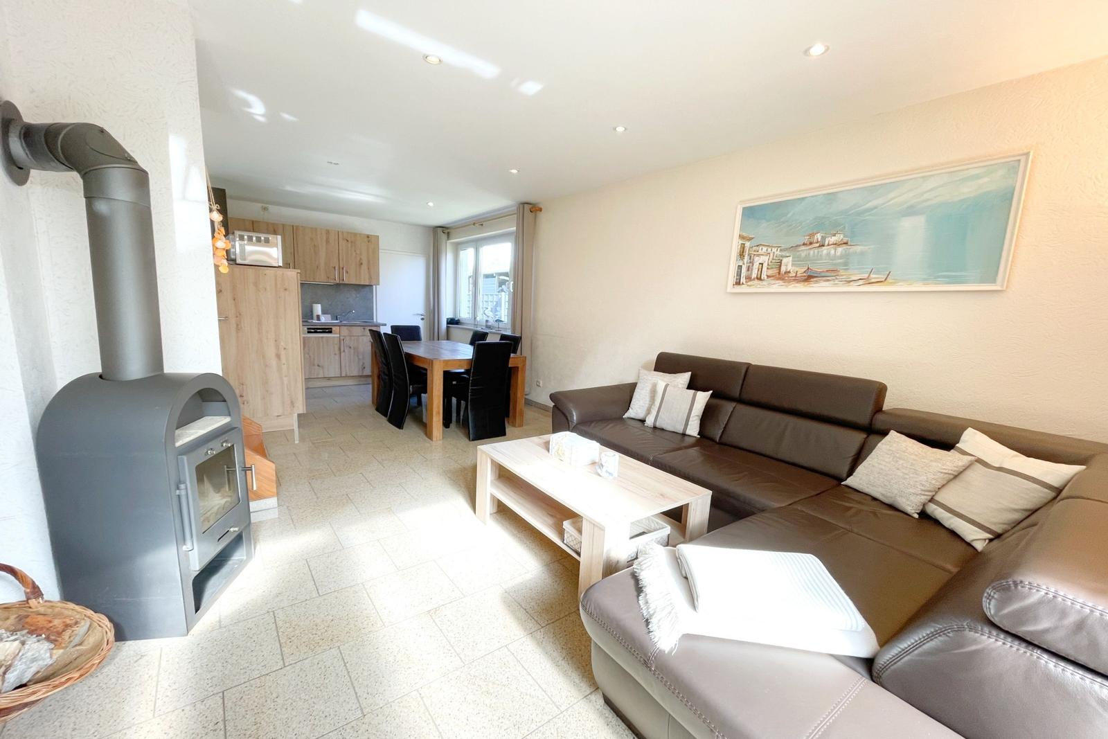 Wohnzimmer mit Sofa, Esstisch und Holzküche. Holzofen und Wandbild sichtbar.