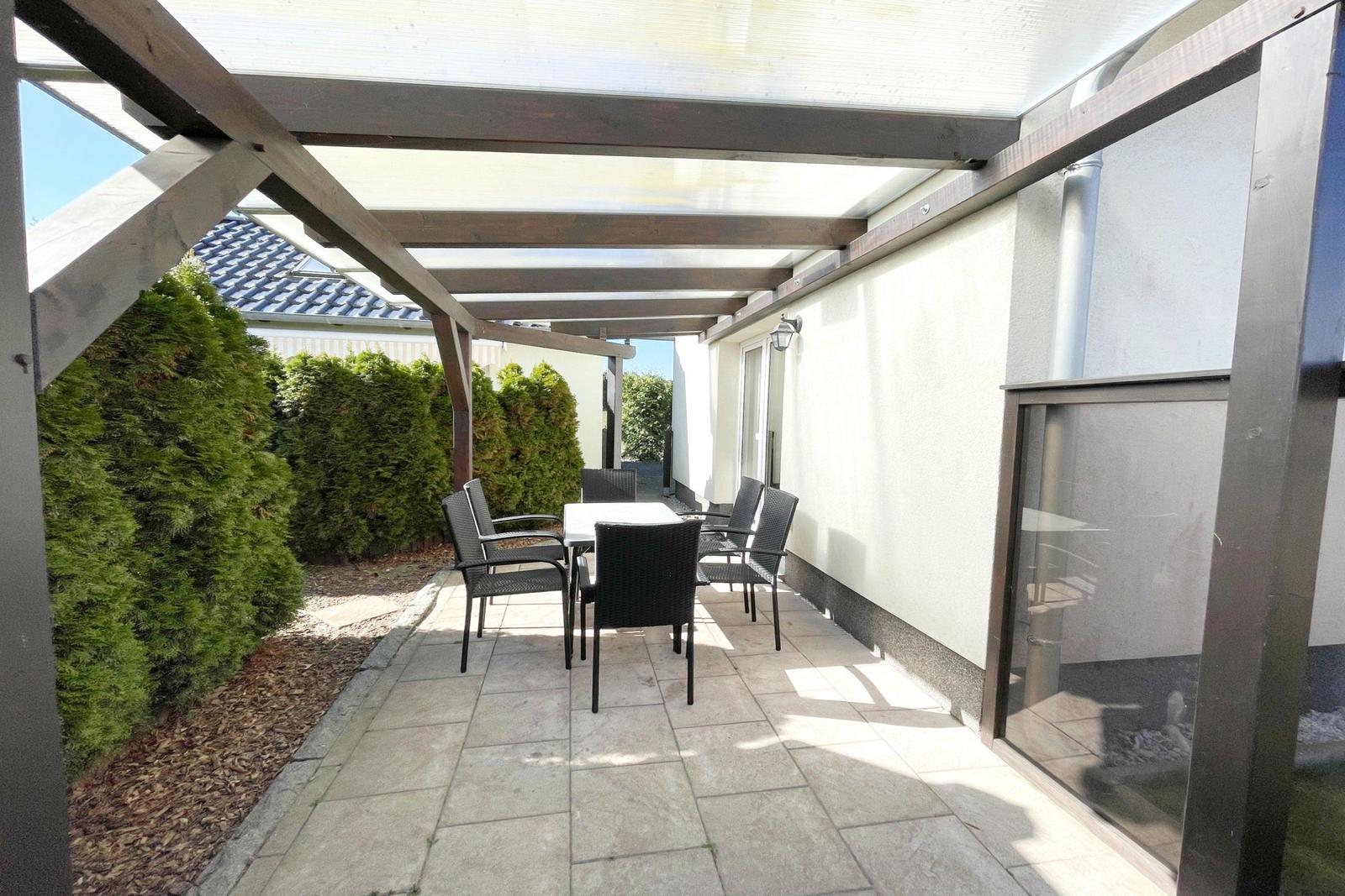 Terrasse mit Tisch und Stühlen unter Dach, umgeben von Hecken und Wand.