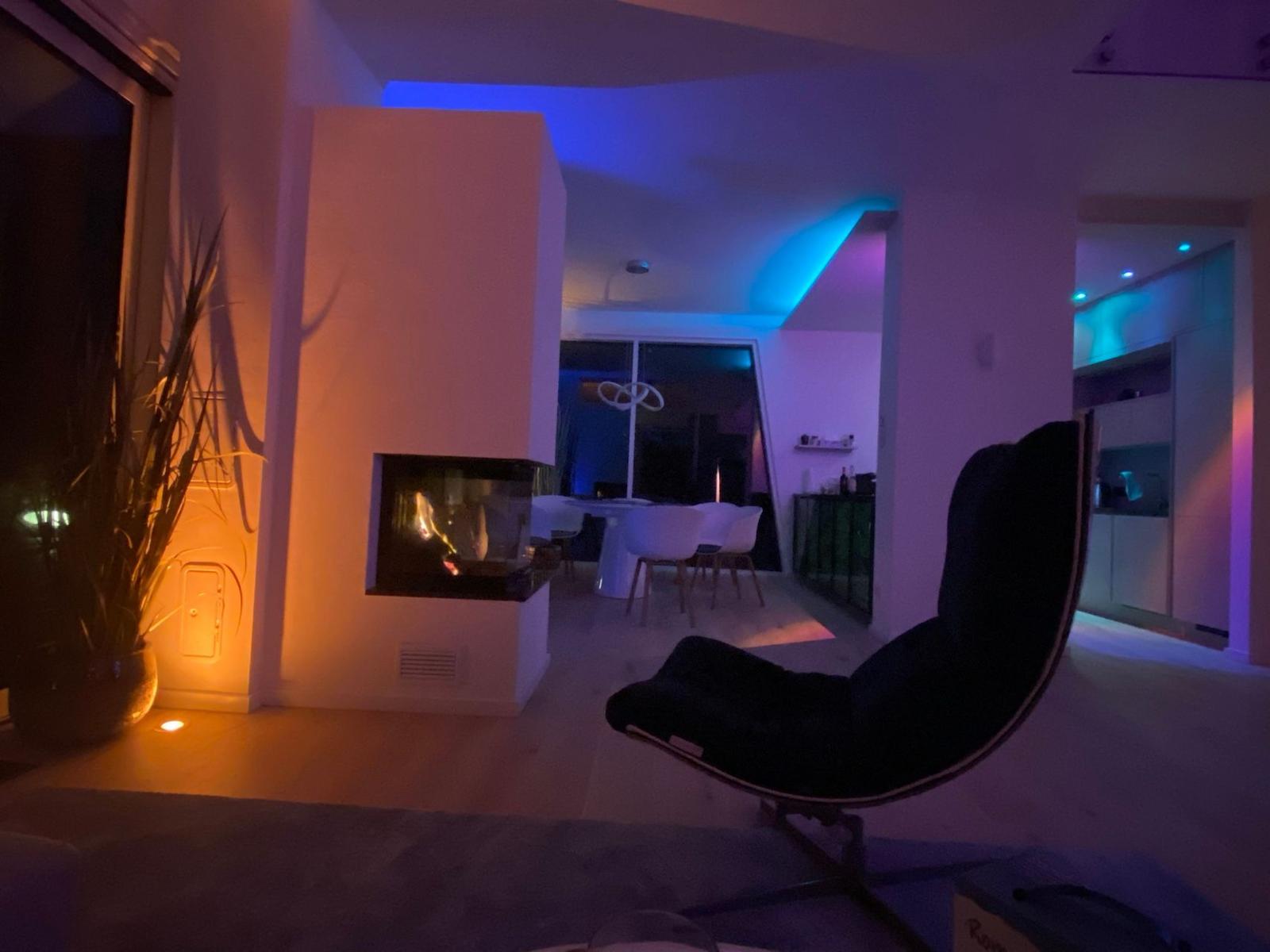 Ein Raum mit Kamin, Sessel und Essbereich. Farbige Beleuchtung und moderne Einrichtung.