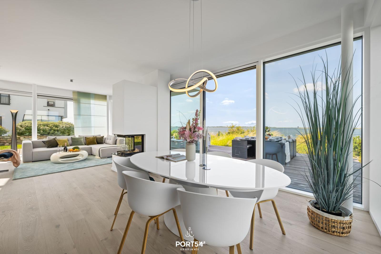 Essbereich mit weißem Tisch und Stühlen. Große Fenster bieten Blick auf Wasser und Terrasse.