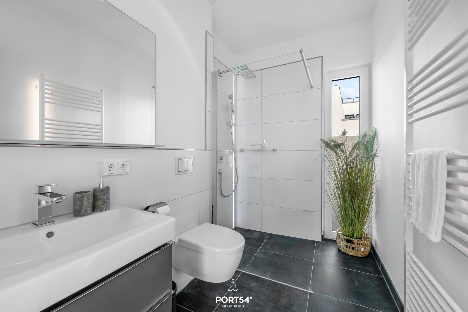 Badezimmer mit Dusche, Waschbecken und Toilette. Weiße Fliesen und dunkler Boden.
