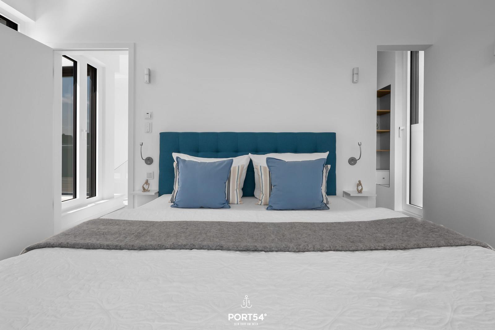 Schlafzimmer mit blauem Kopfteil, weißen Wänden und Fenstern.