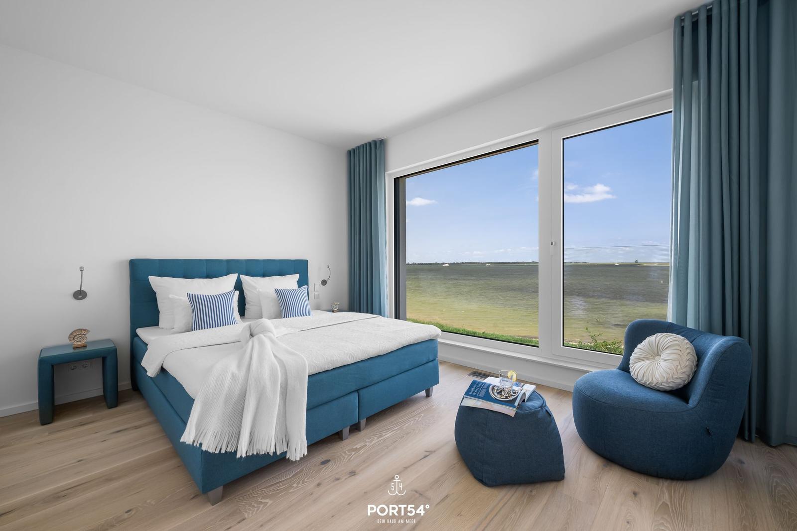Schlafzimmer mit Blick auf Wasser, blaues Bett und Sessel.