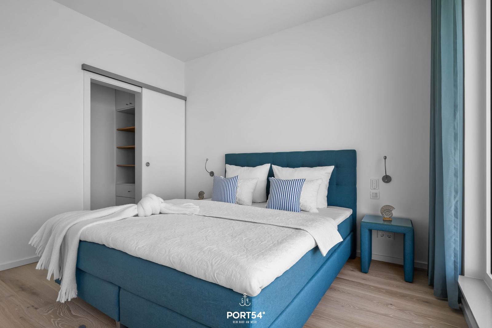 Schlafzimmer mit blauem Bett, Nachttisch und Gardinen.