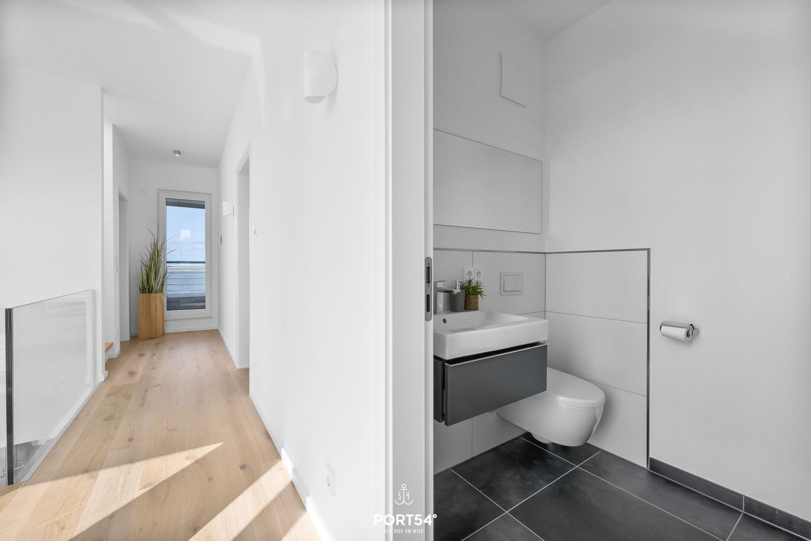 Flur mit Holzboden und Blick auf Balkon. Nebenbei ist das Badezimmer mit Toilette und Waschbecken.