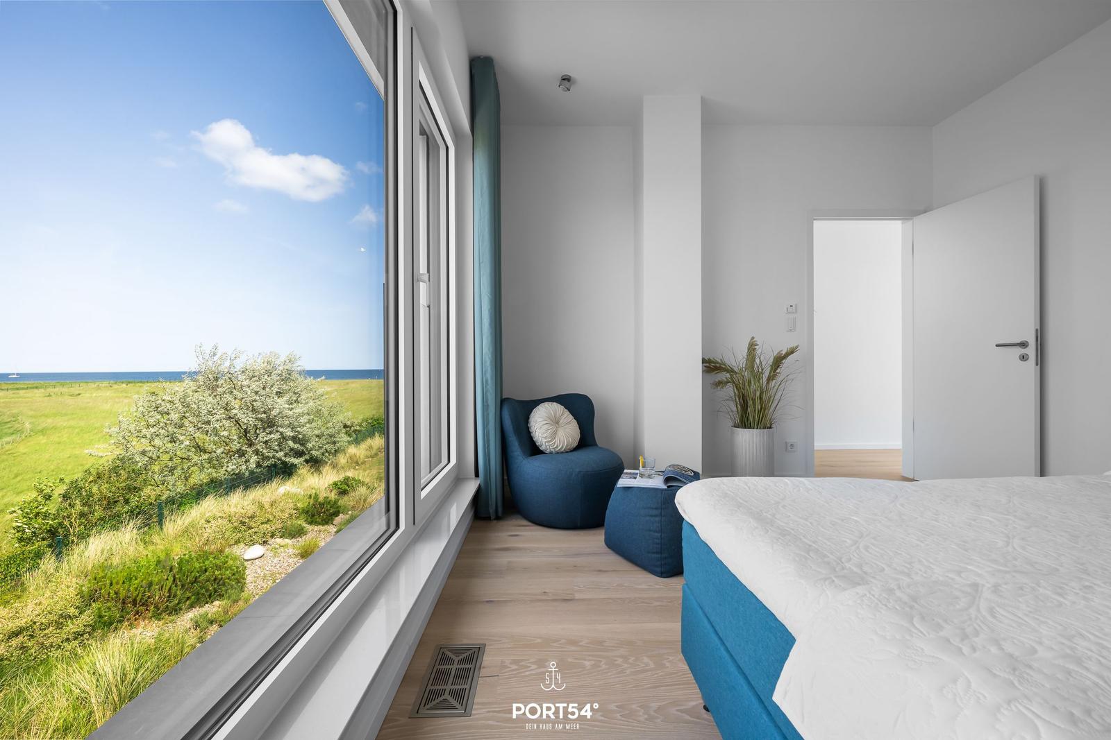 Schlafzimmer mit Blick auf Grasland und Meer. Blauer Sessel und Bett mit weißer Bettwäsche.