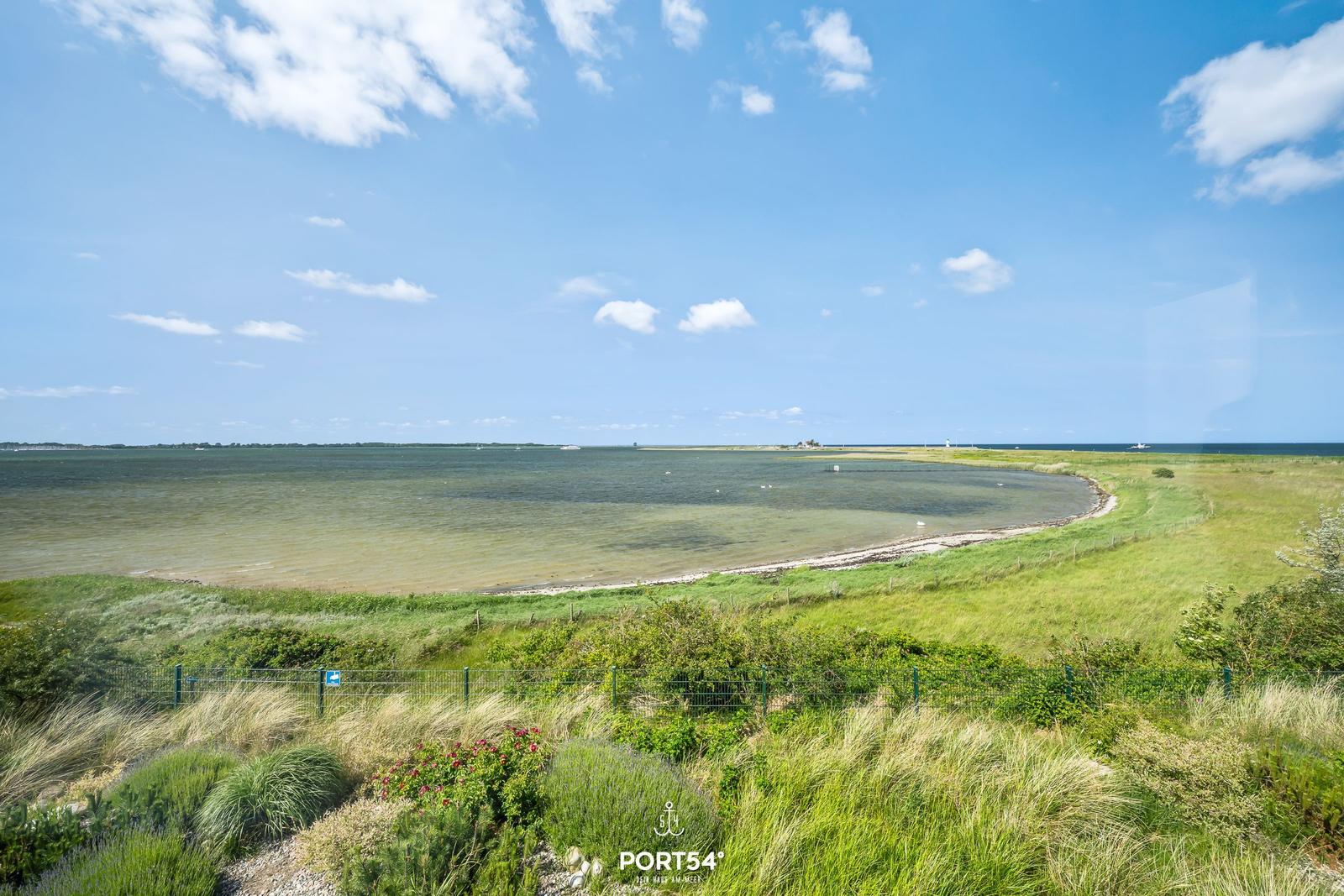 Weitläufiger Blick auf Küstenlandschaft mit Grünflächen und Wasser unter blauem Himmel.