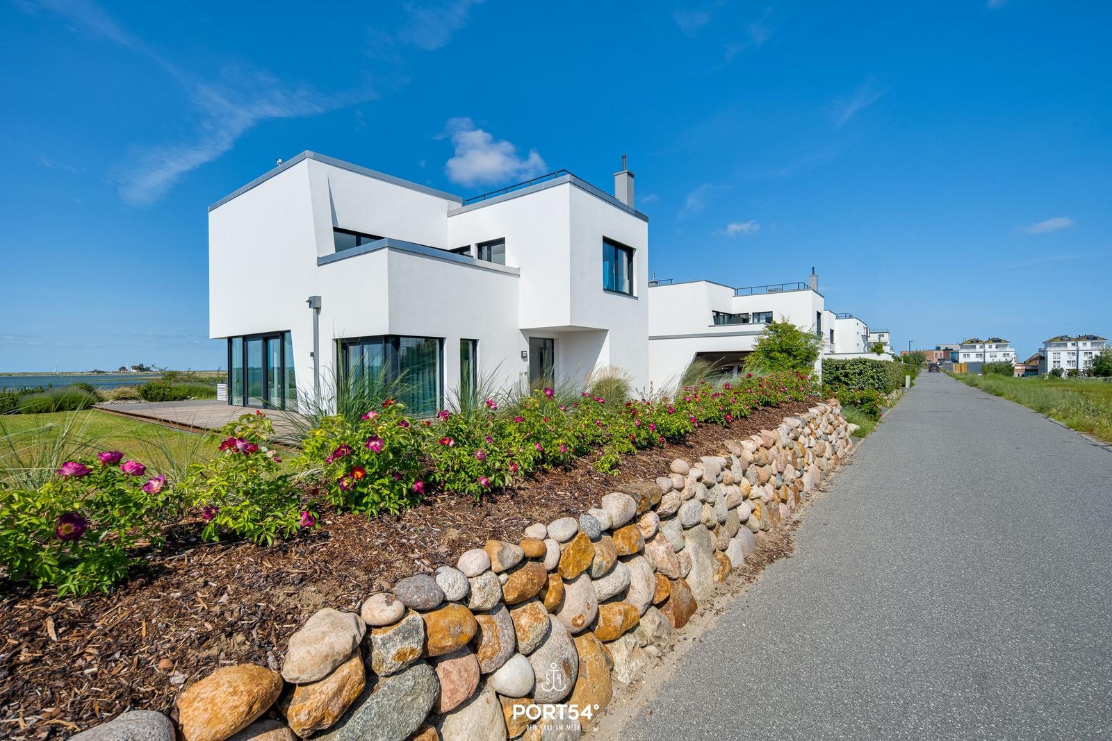 Moderne weiße Häuser am Wasser mit Steinmauer und Blumenbeeten.