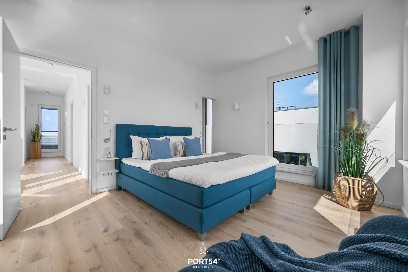 Schlafzimmer mit blauem Bett, Holzboden und großem Fenster mit Blick.