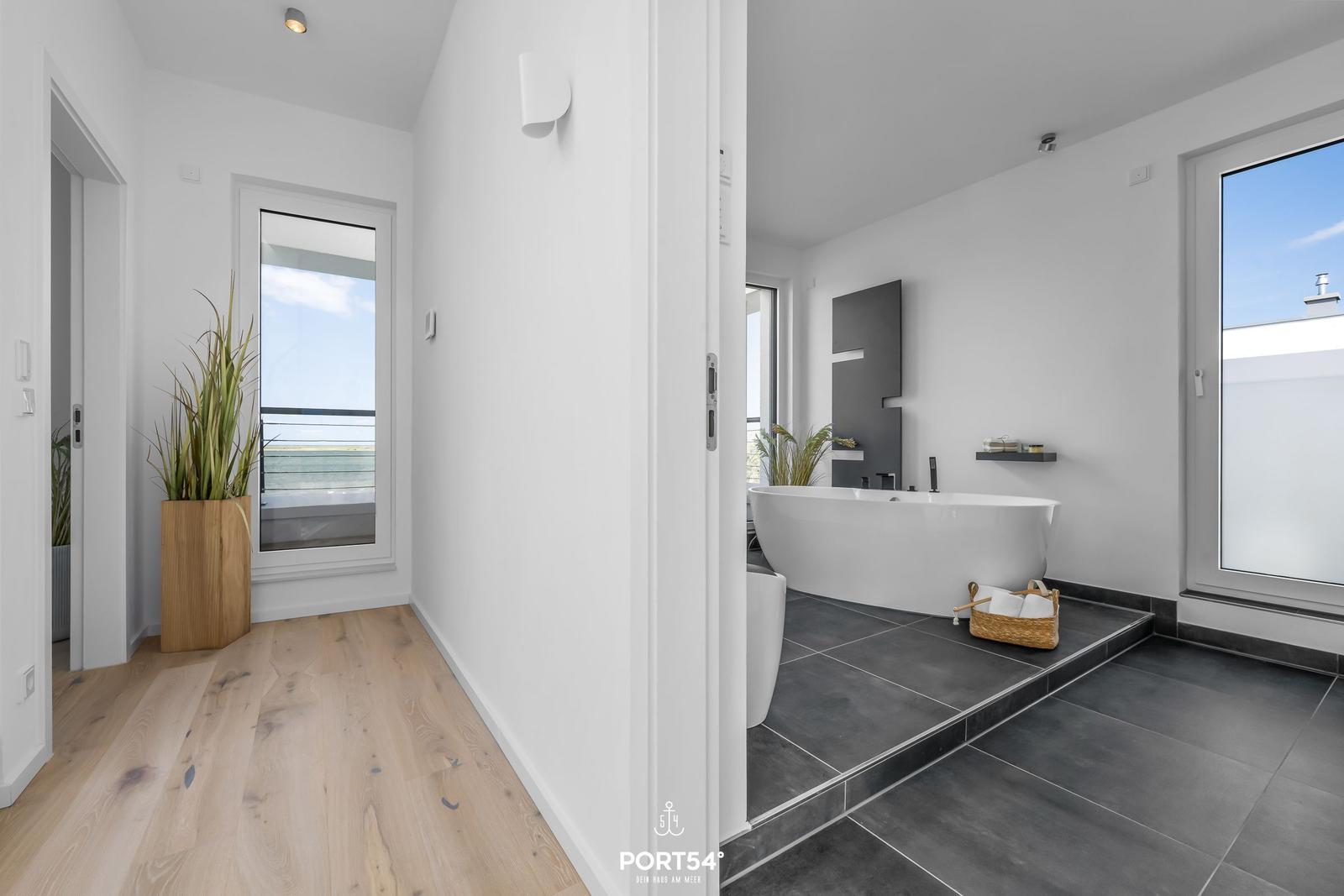 Badezimmer mit freistehender Badewanne und großem Fenster. Holzfußboden und Pflanze im Flur.