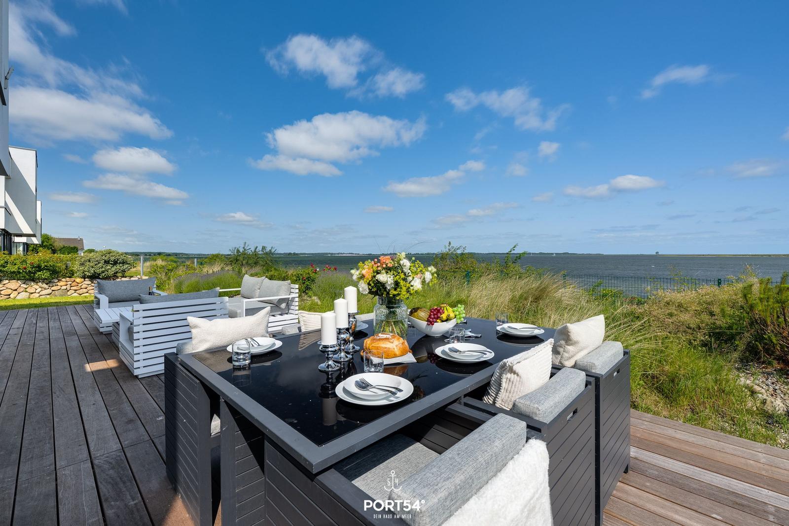 Terrasse mit Tisch und Sitzgelegenheiten. Blick auf Wasser und Himmel.